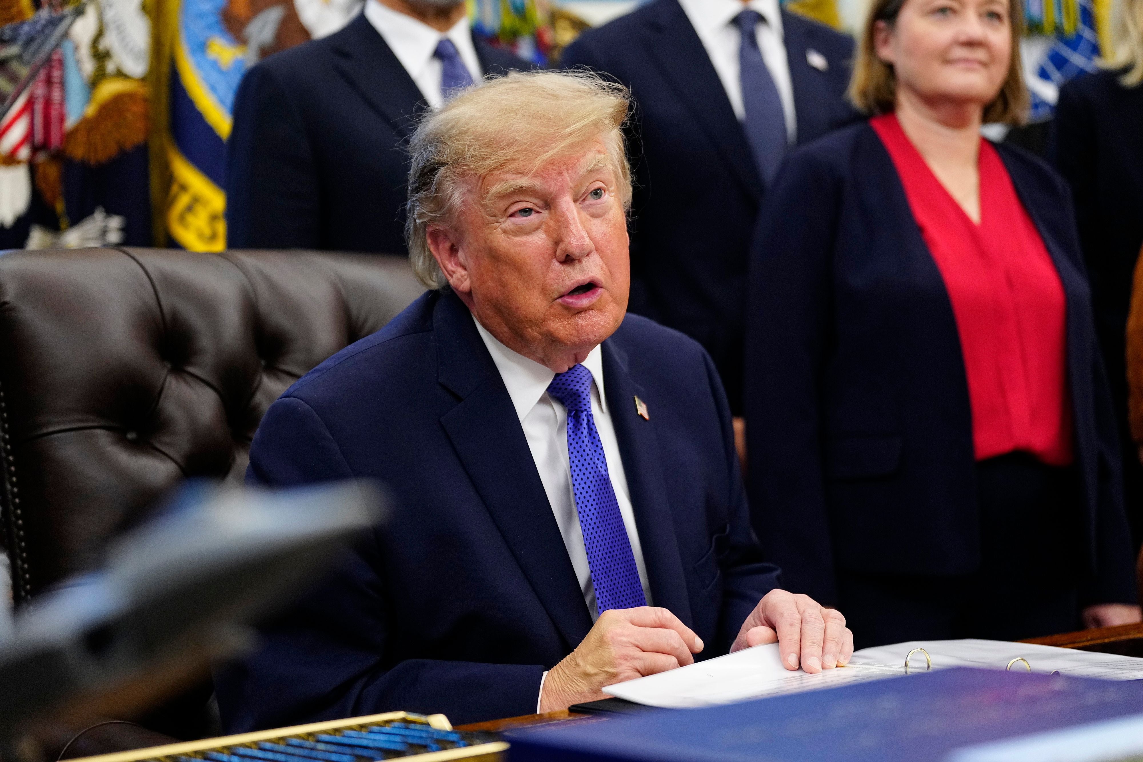 Ein Mann im Anzug spricht am Schreibtisch im Oval Office, mehrere Personen stehen hinter ihm.