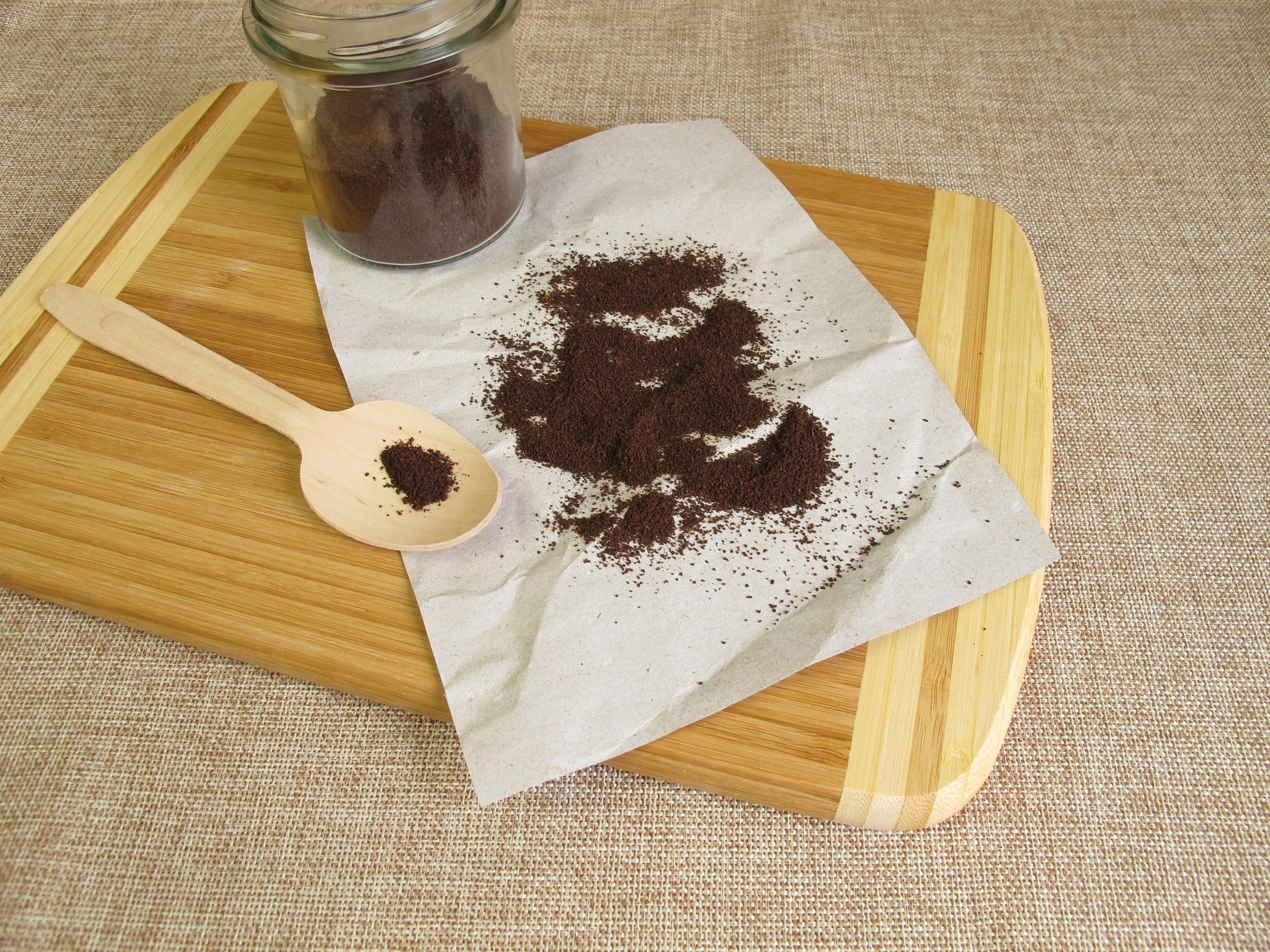 Getrockneter Kaffeesatz auf Papier, daneben ein Glas und ein Holzlöffel auf einem Holzbrett.