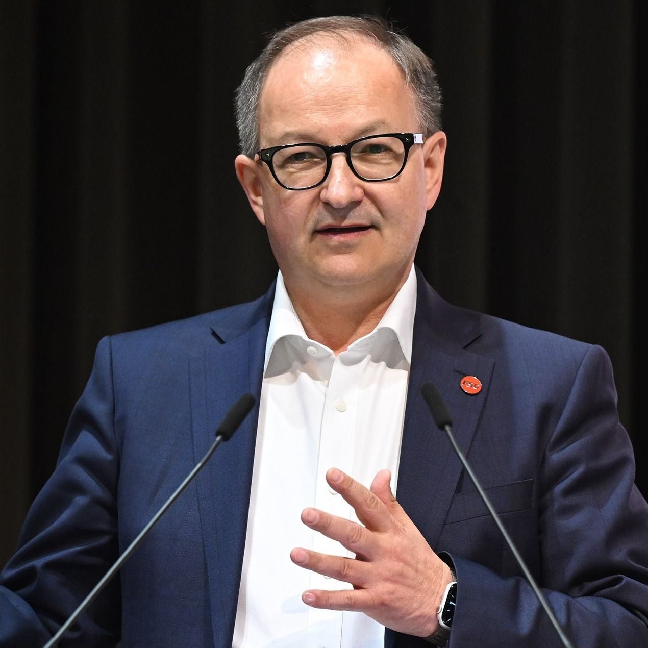Ein Mann mit Brille spricht am Rednerpult und trägt einen dunklen Anzug mit rotem Anstecker.