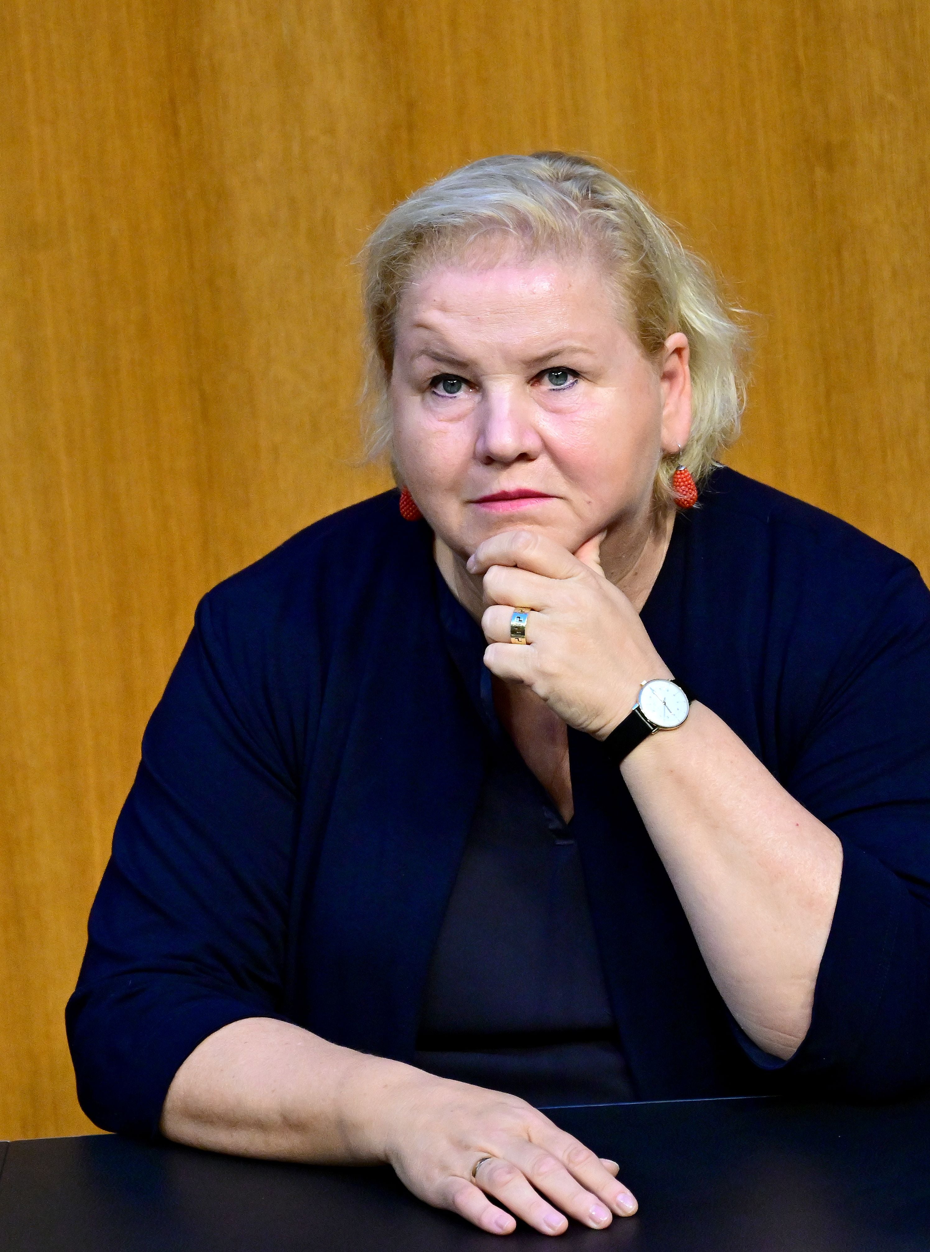 Eine nachdenklich blickende Frau sitzt vor einer Holzwand an einem Tisch.