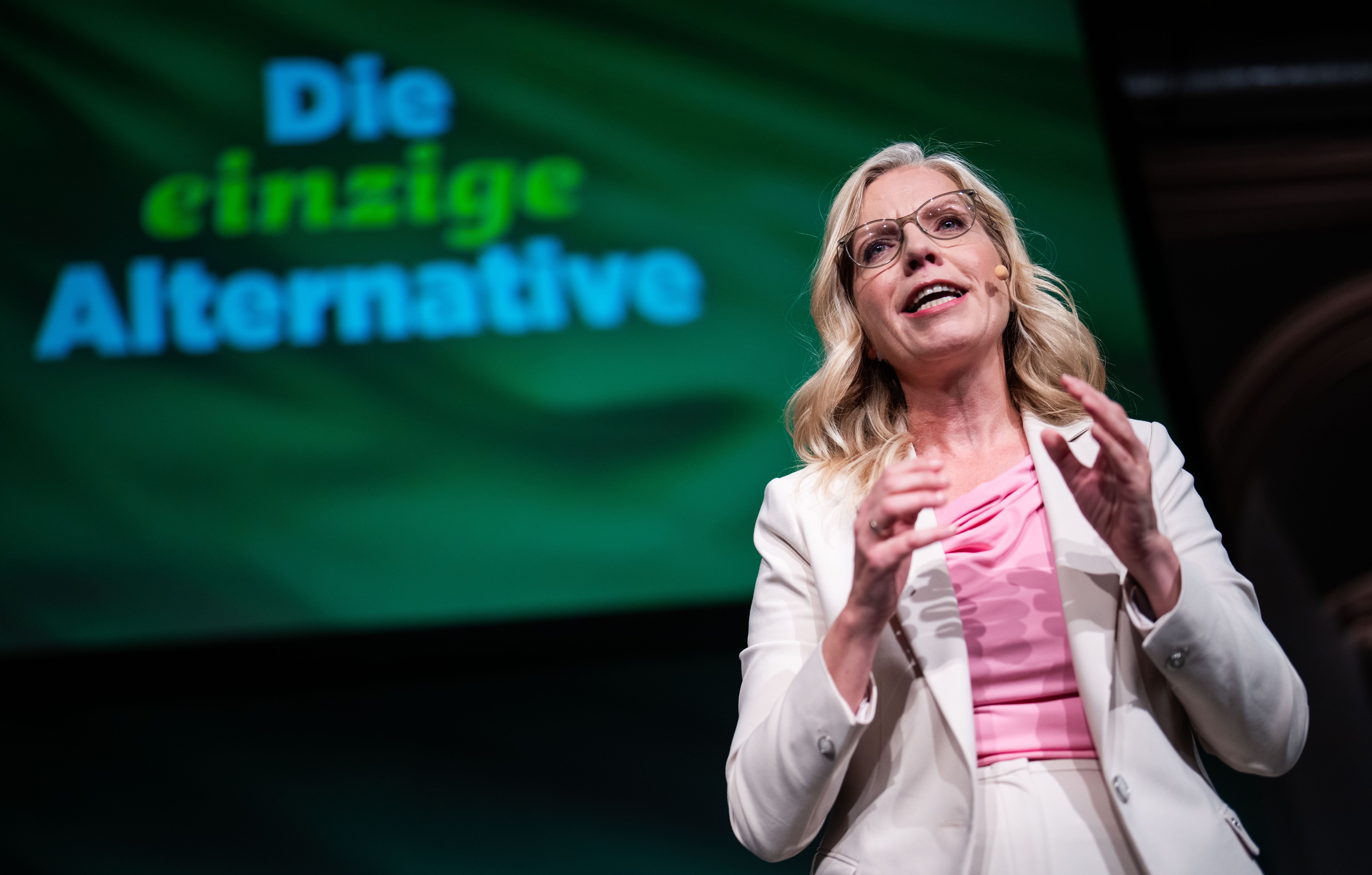 Frau in weißem Anzug spricht auf Bühne vor grünem Hintergrund mit Text „Die einzige Alternative“.