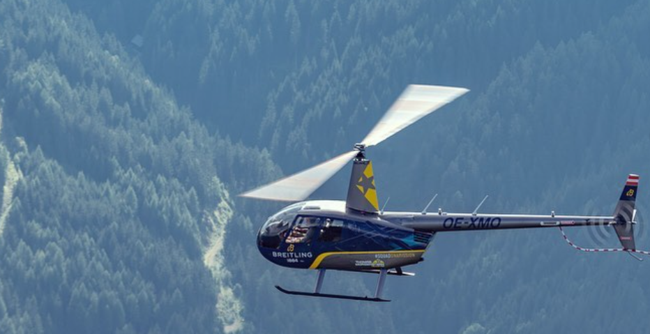 Ein dunkelblauer Helikopter fliegt vor bewaldeten Berghängen.
