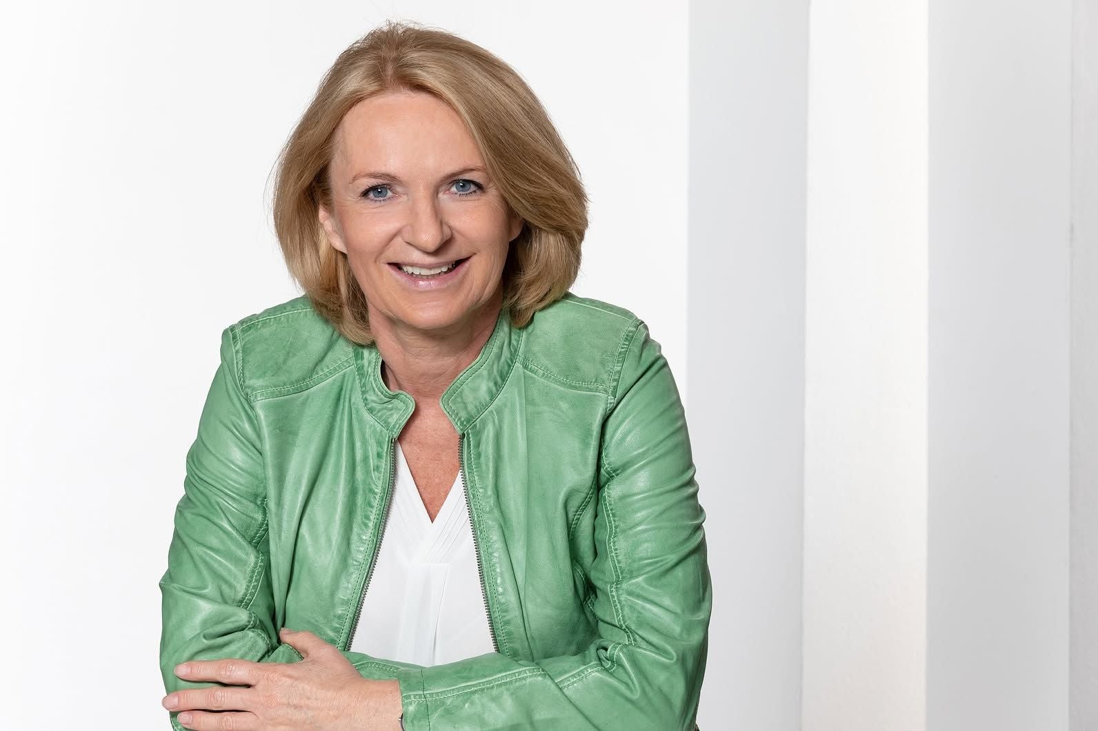 Frau mit blonden Haaren und grüner Lederjacke lächelt in die Kamera vor weißem Hintergrund.
