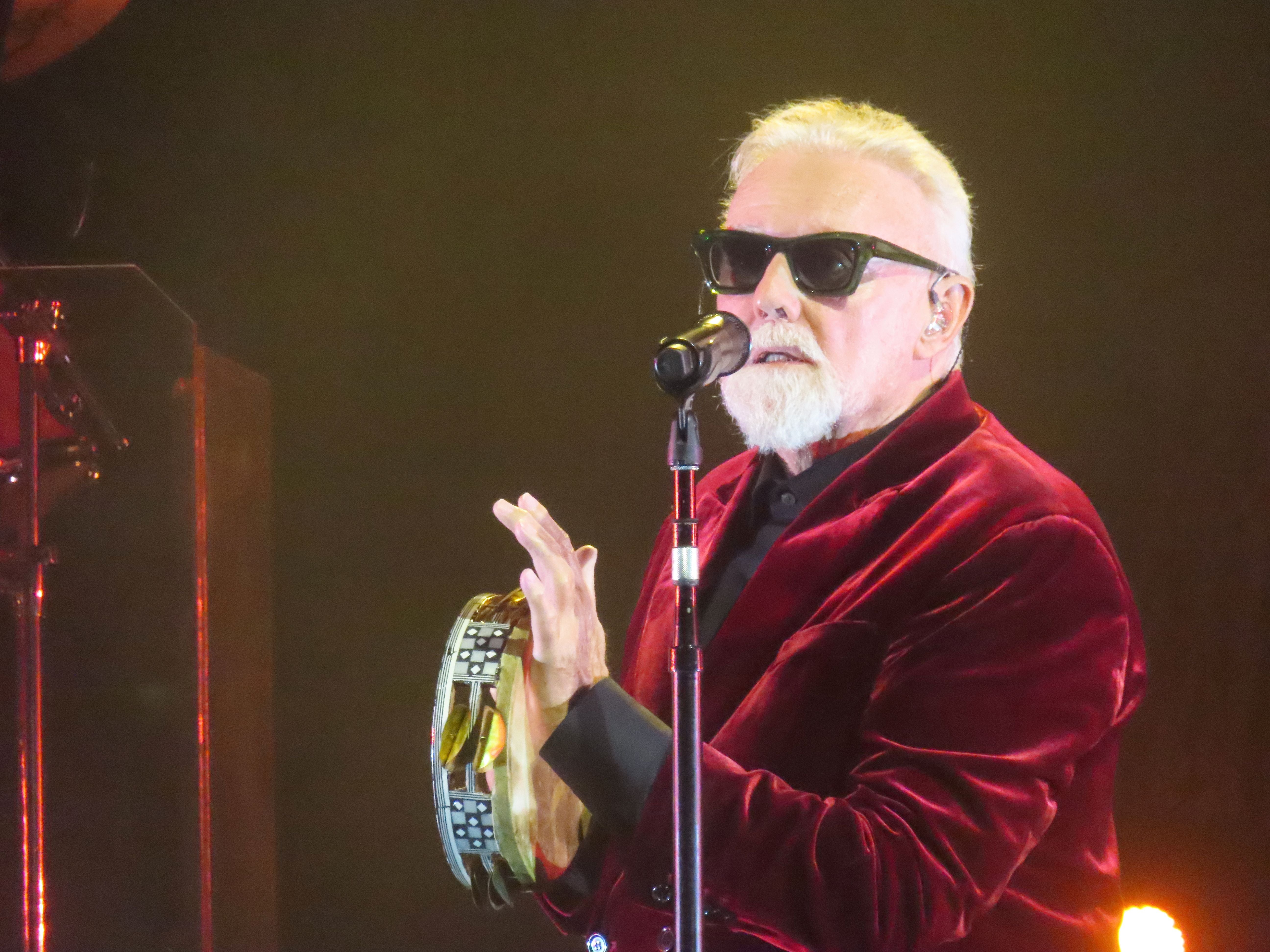 Ein älterer Mann im roten Samtjackett singt mit Tamburin und Sonnenbrille auf der Bühne.