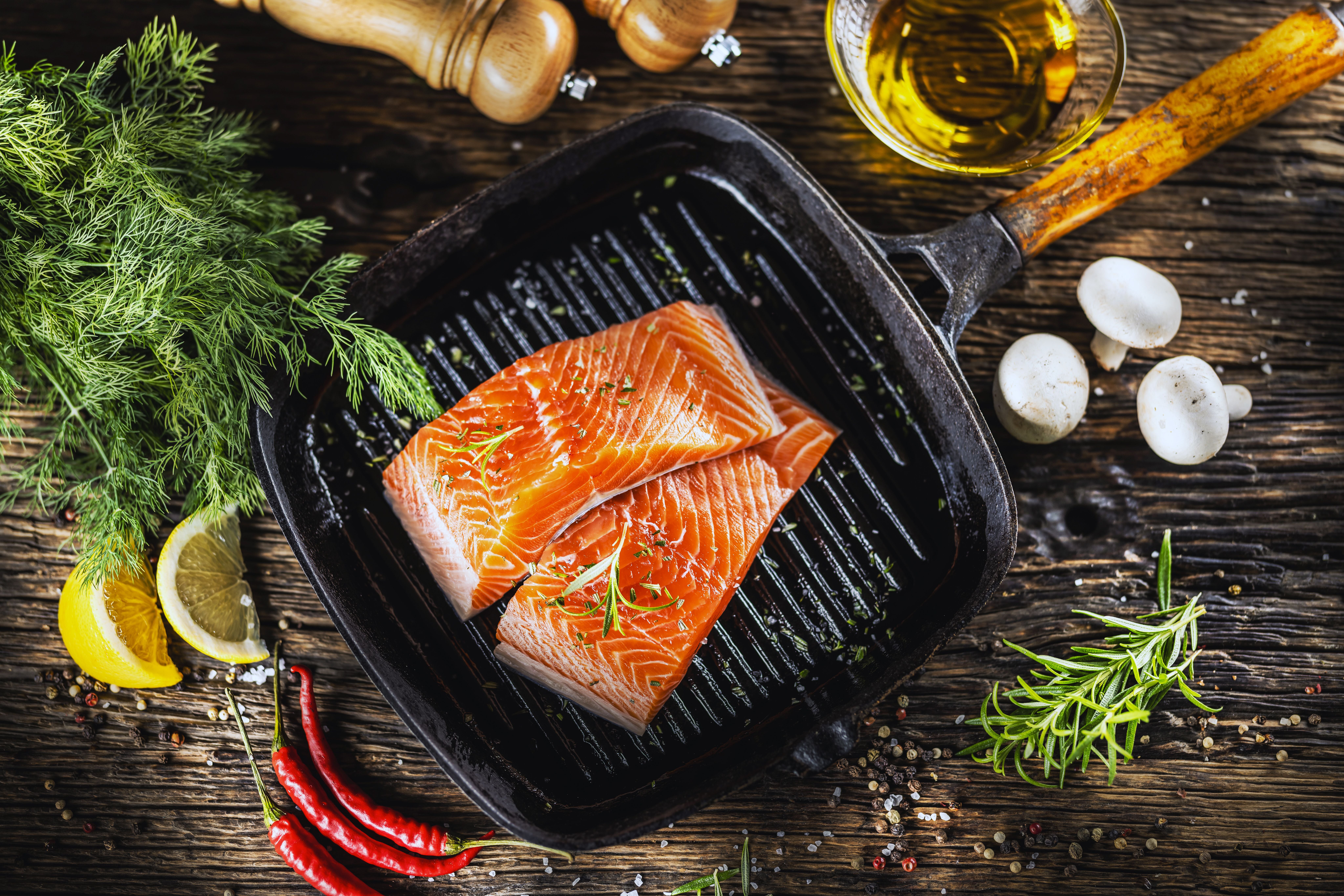 Zwei rohe Lachsfilets in einer Grillpfanne, umgeben von Kräutern, Zitrone, Chili, Pilzen und Öl.