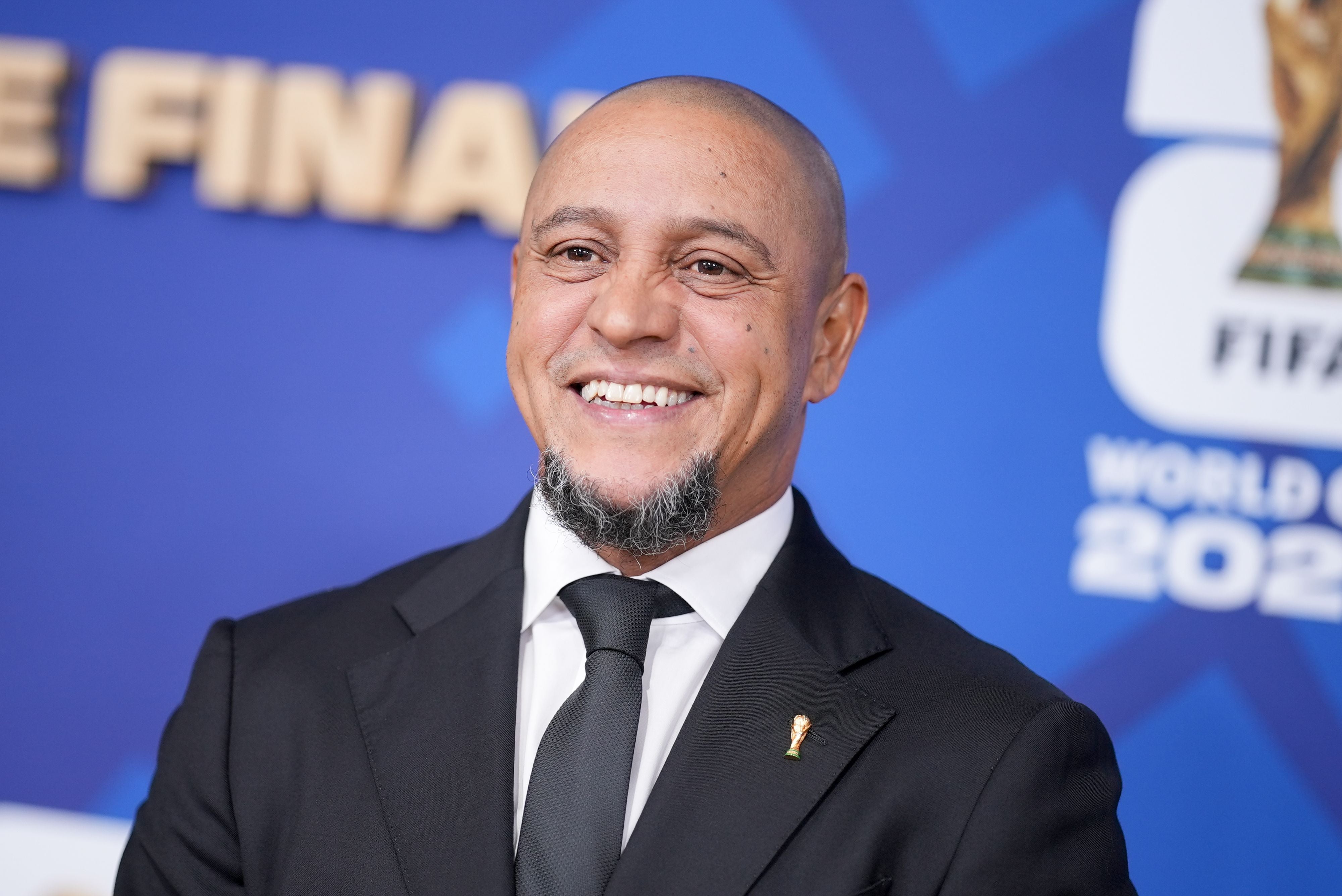 Ein lächelnder Mann im Anzug steht vor einer blauen FIFA World Cup 2026 Hintergrundwand.