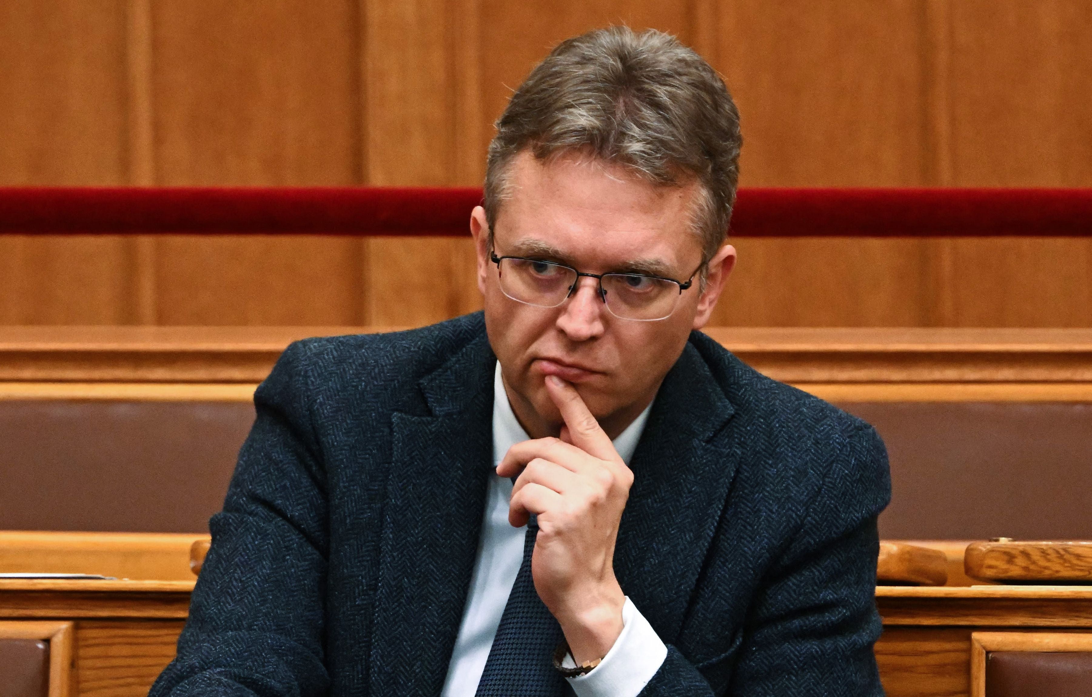 Ein Mann mit Brille und Anzug sitzt nachdenklich in einem Holzsaal, den Finger am Kinn.