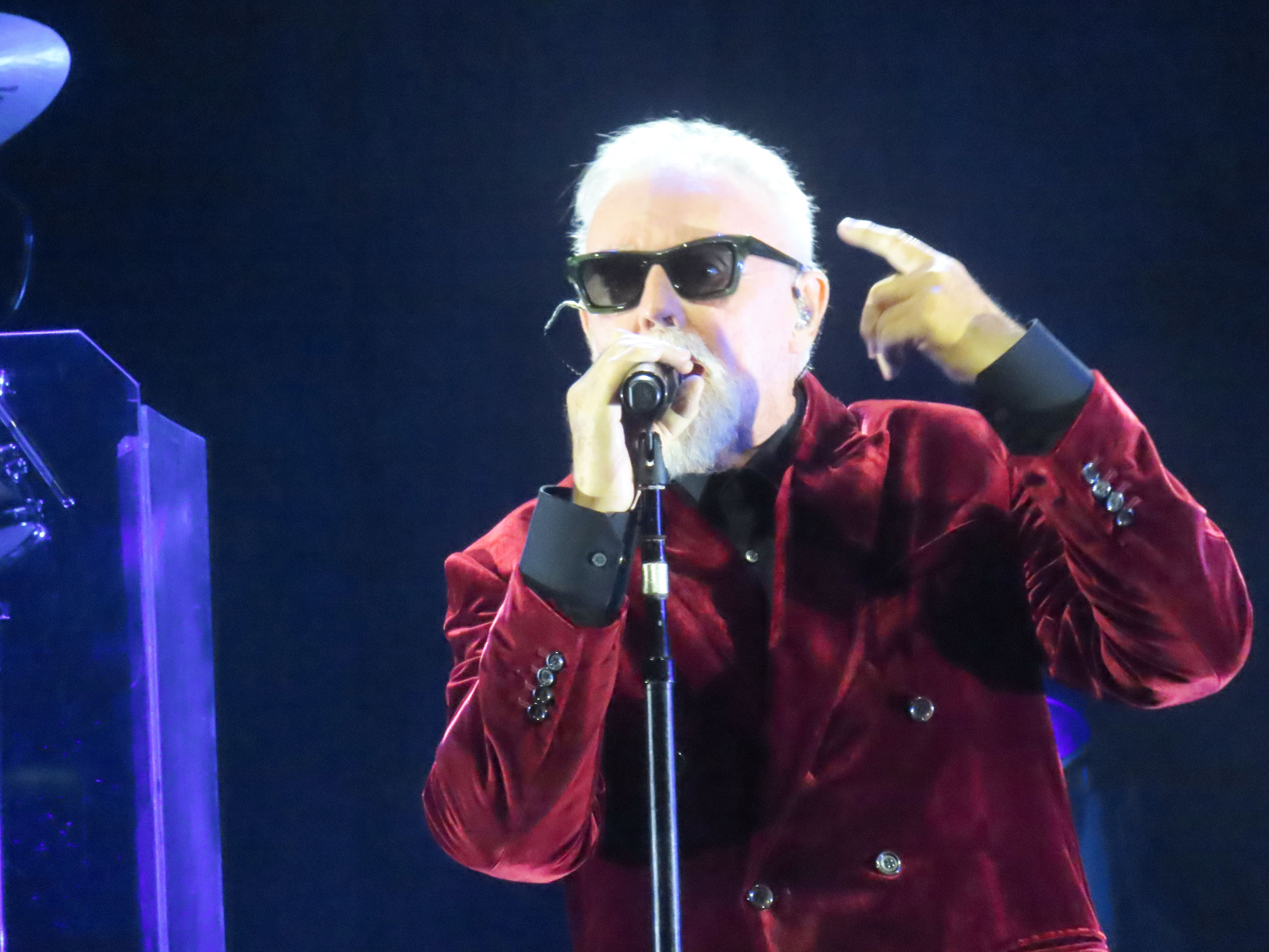 Mann mit Sonnenbrille und rotem Samtjackett singt auf einer Bühne ins Mikrofon.