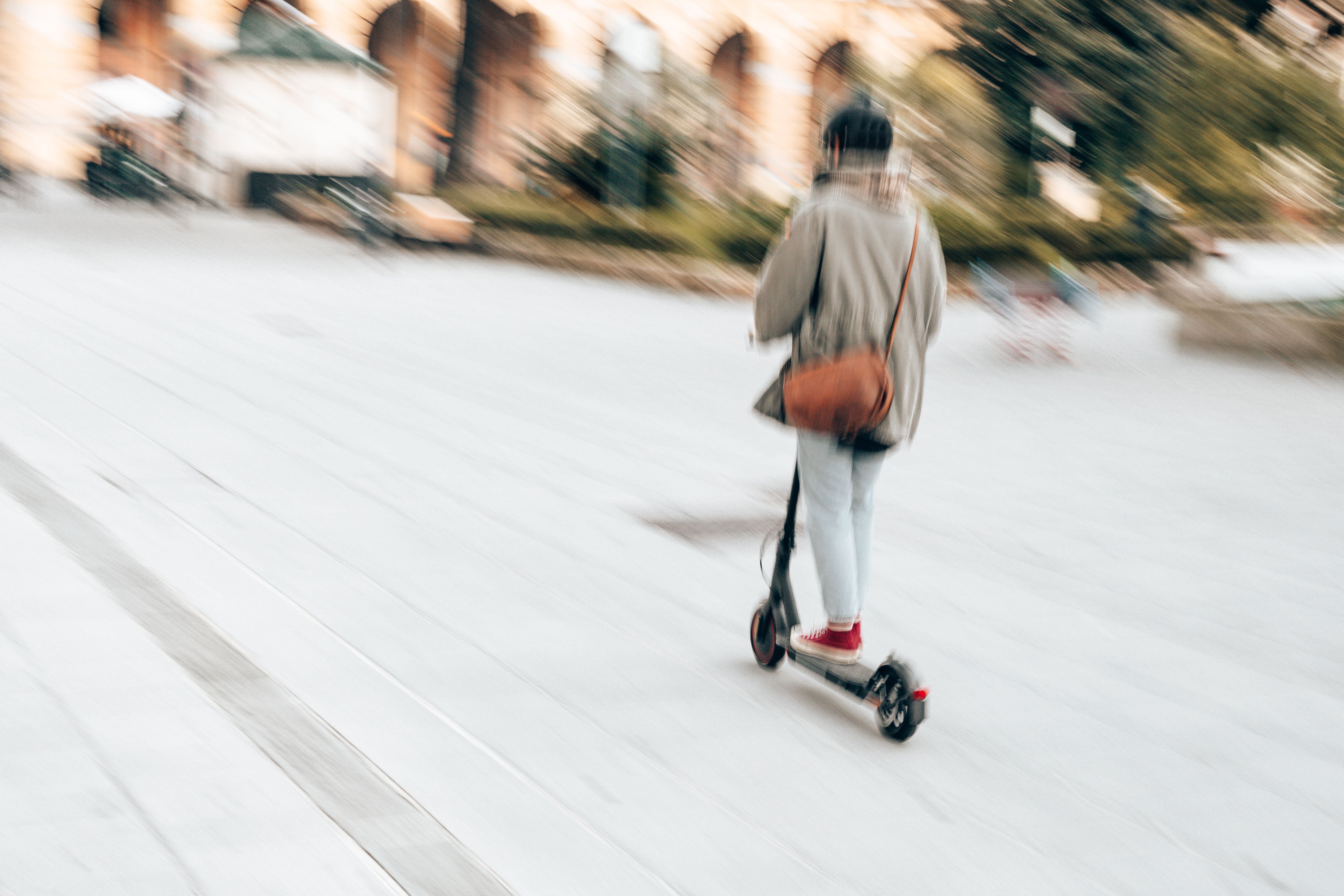 Verschwommene Frau mit Maske fährt auf einem E-Scooter durch eine Stadt.
