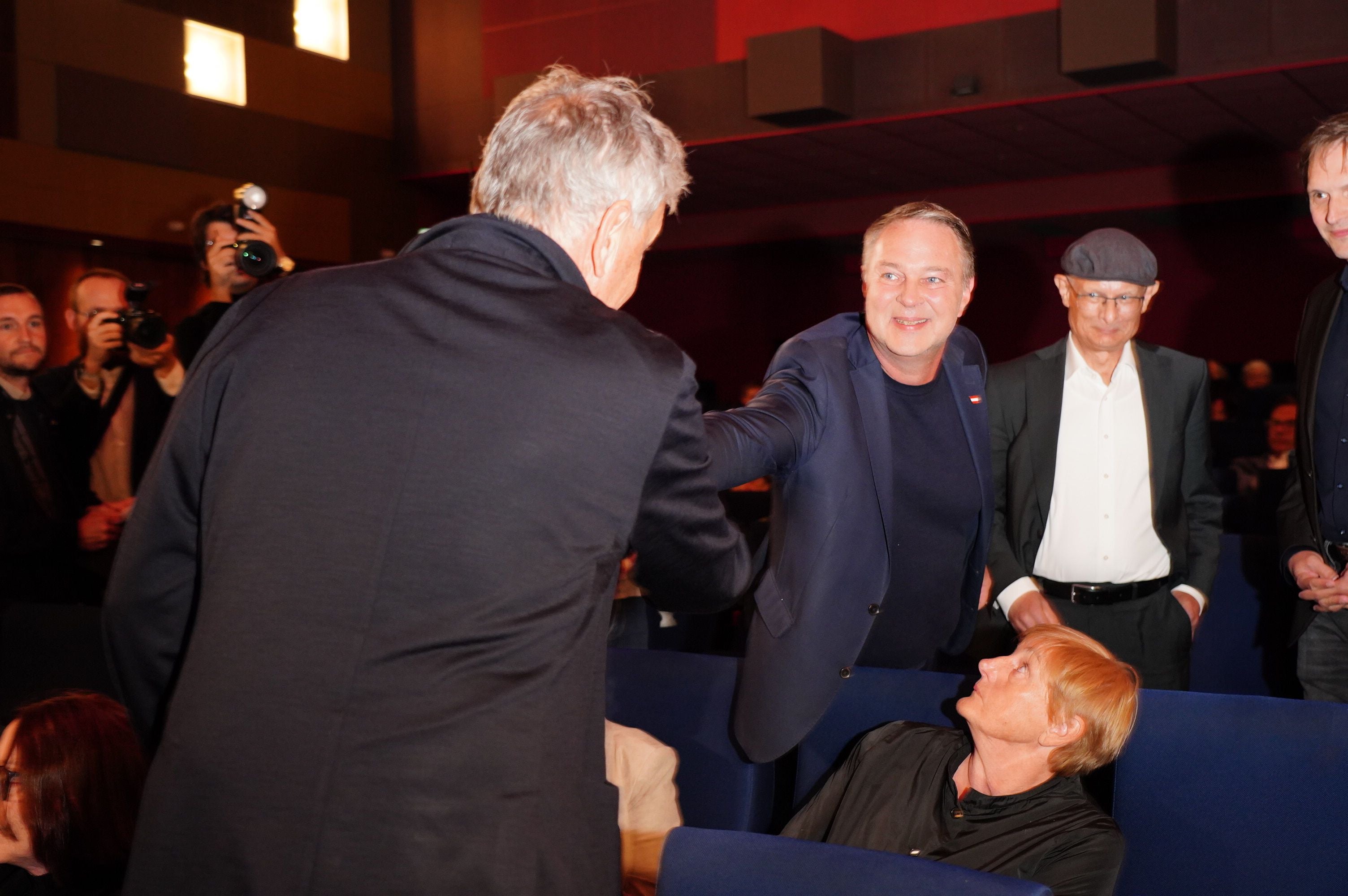 Zwei Männer geben sich in einem Saal die Hand, umgeben von weiteren Personen.