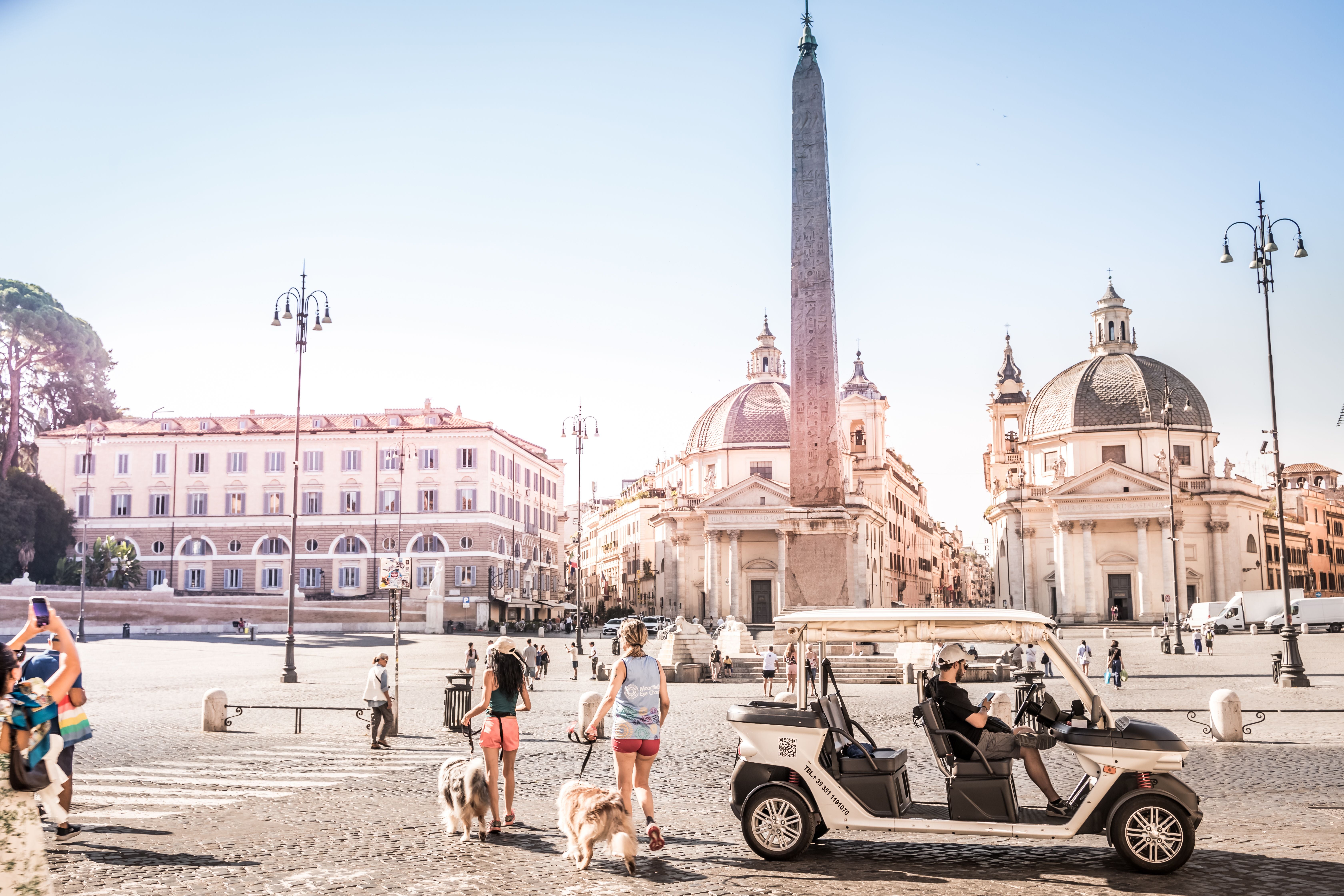 Menschen und Hunde spazieren auf der Piazza del Popolo in Rom, im Hintergrund Kirchen und Obelisk.