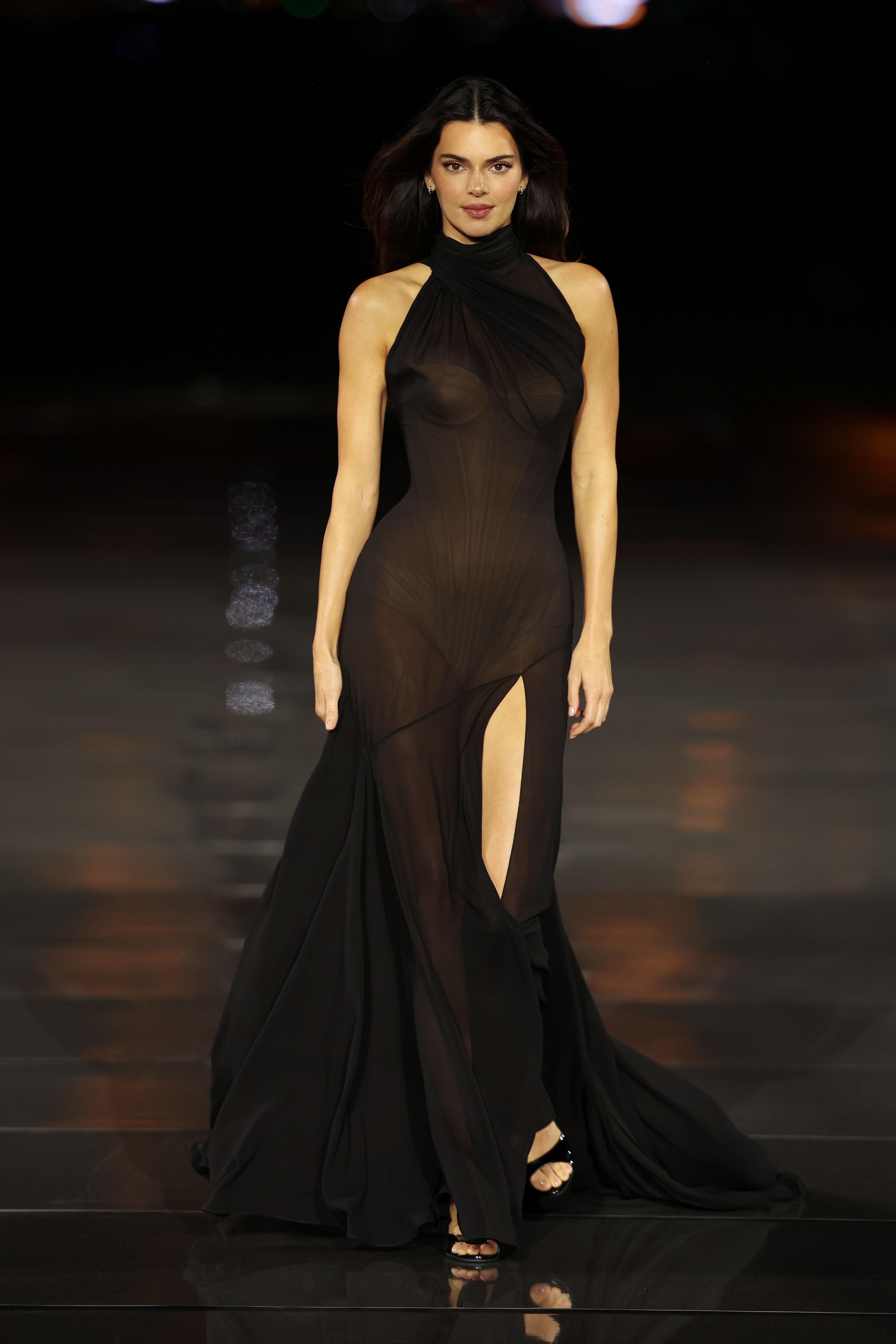Model läuft in einem langen, transparenten schwarzen Kleid mit Beinschlitz über den Laufsteg.