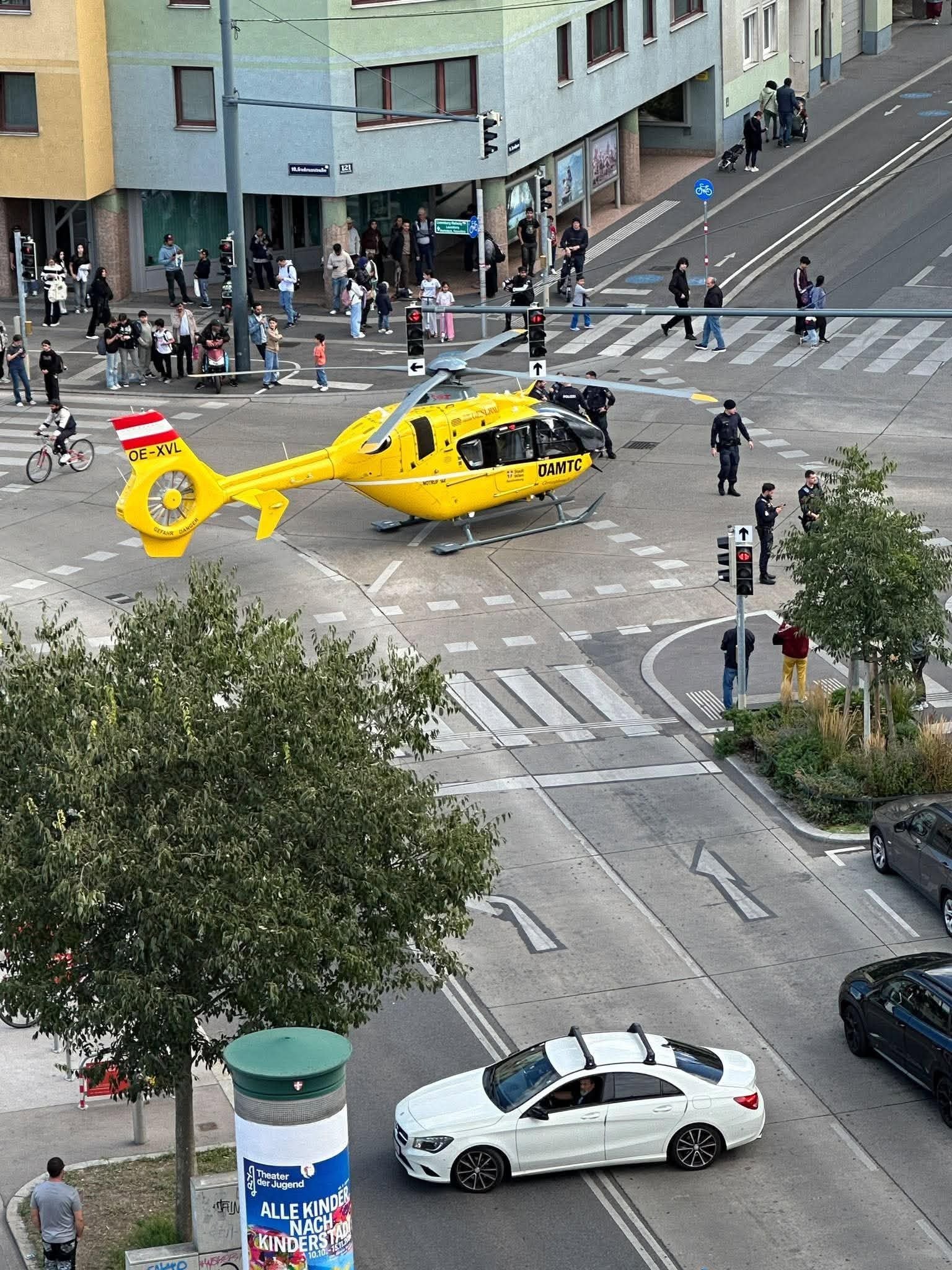 Ein gelber Rettungshubschrauber landet an einer Kreuzung, Polizei und Schaulustige stehen drumherum.