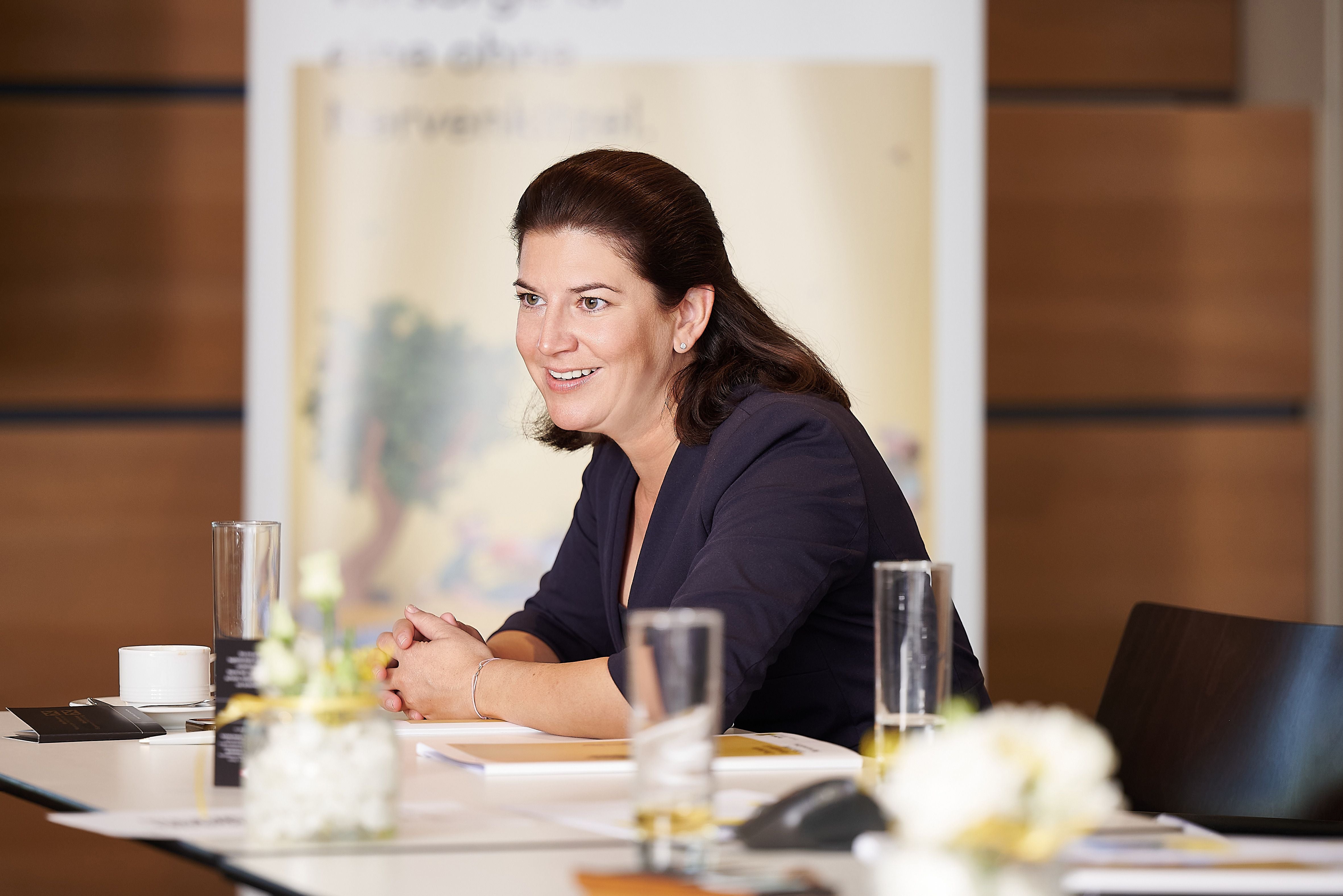Frau mit dunklen Haaren sitzt lächelnd an einem Konferenztisch mit Getränken und Unterlagen.