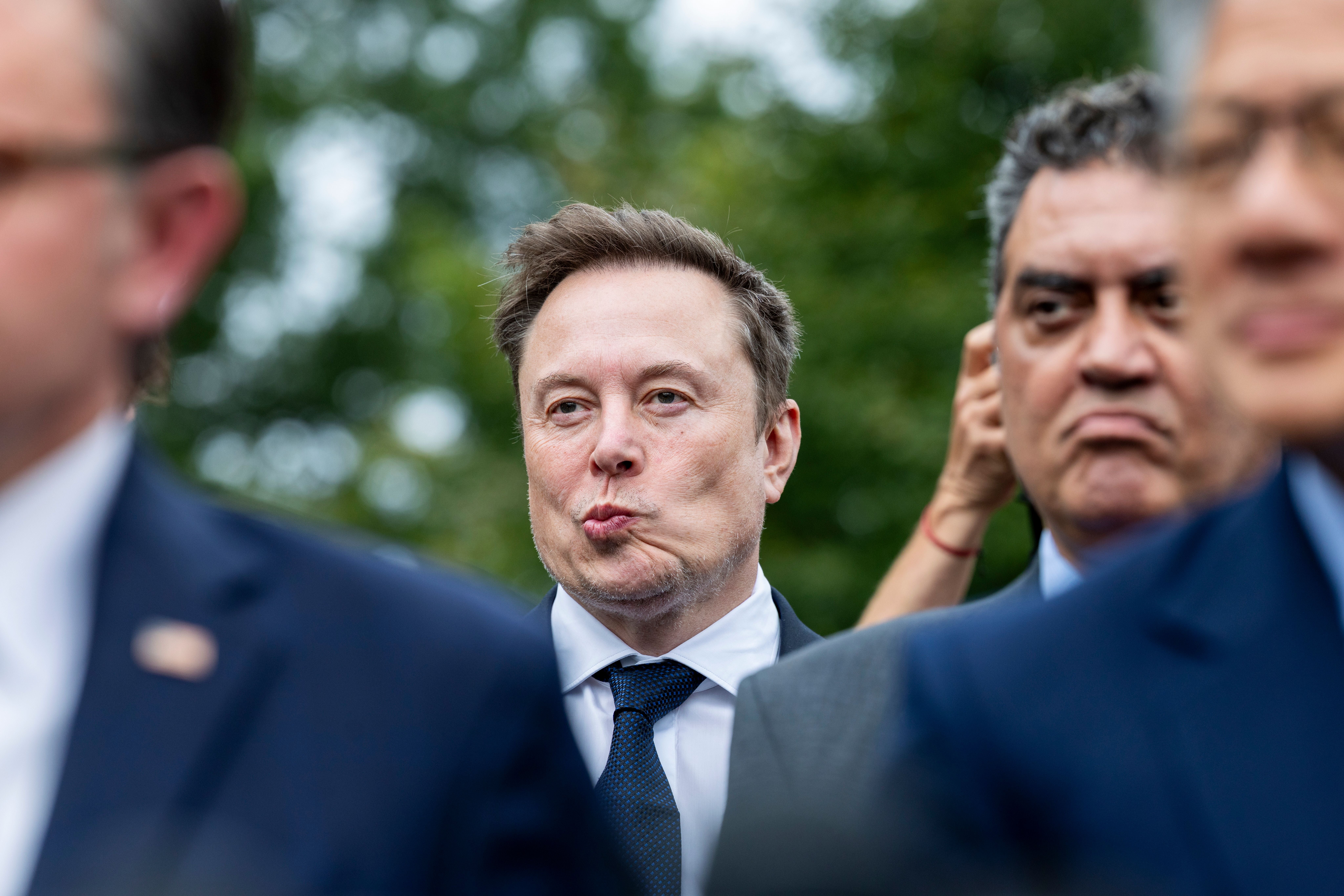 Ein Mann in Anzug macht ein schmollendes Gesicht, weitere Männer stehen im Hintergrund.