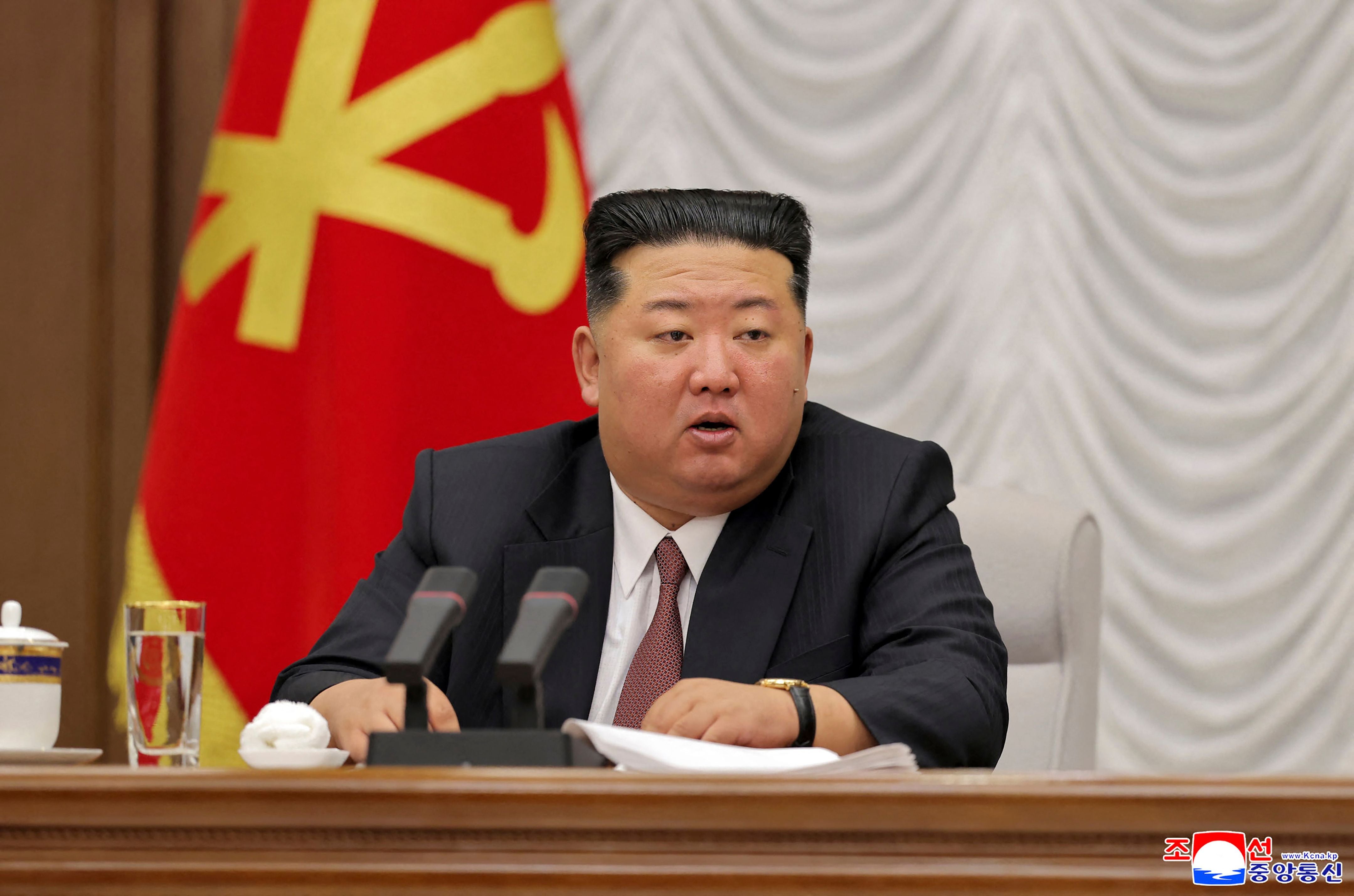 Kim Jong Un sitzt im Anzug an einem Tisch mit Mikrofonen, im Hintergrund eine rote Fahne.