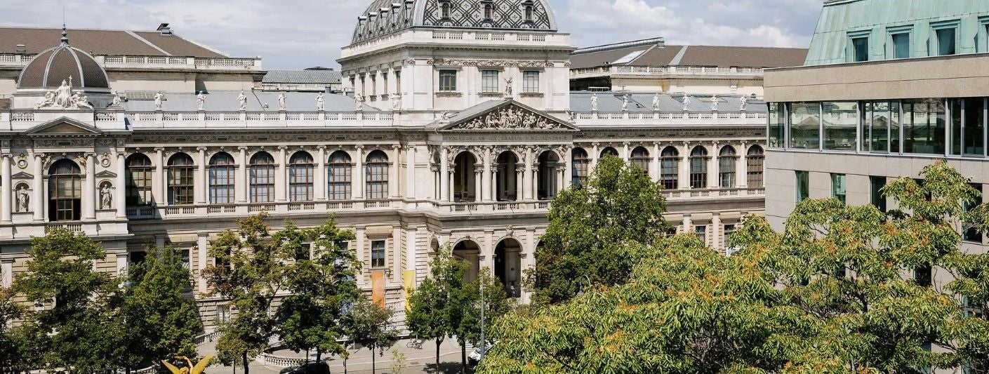 Hauptgebäude der Universität Wien hinter grünen Bäumen an einem sonnigen Tag.