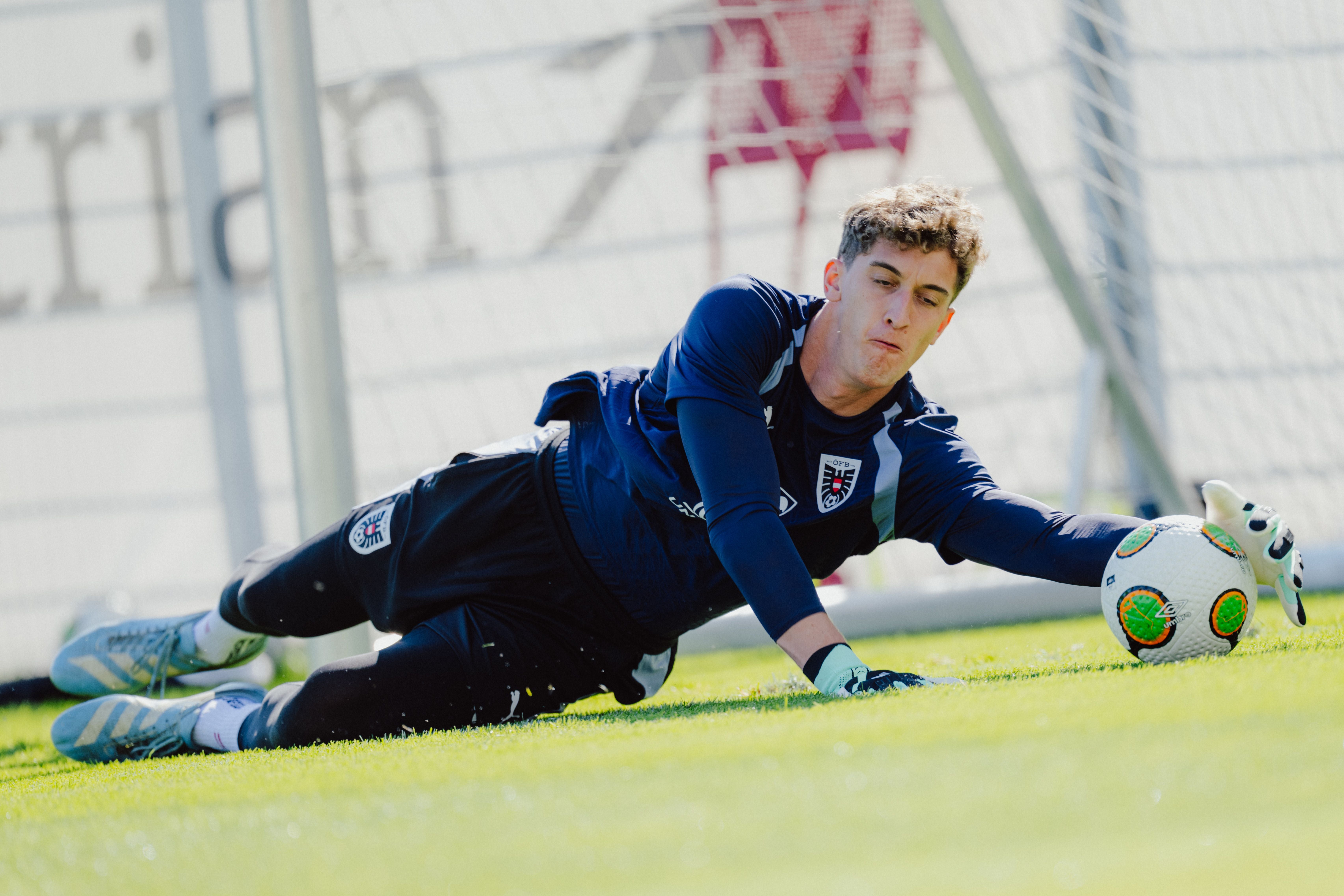 Ein Torwart in dunkelblauer Kleidung hält beim Training einen Fußball am Boden fest.