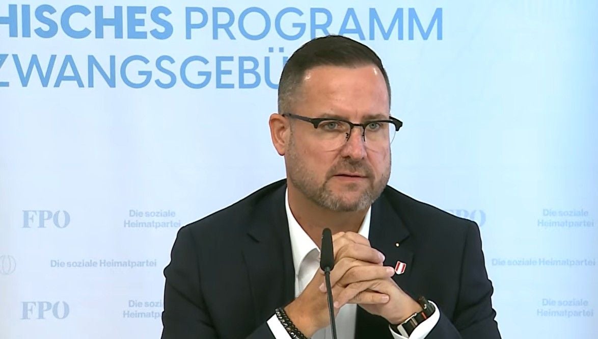 Ein Mann mit Brille spricht bei einer Pressekonferenz, Hintergrund mit blauem Text.