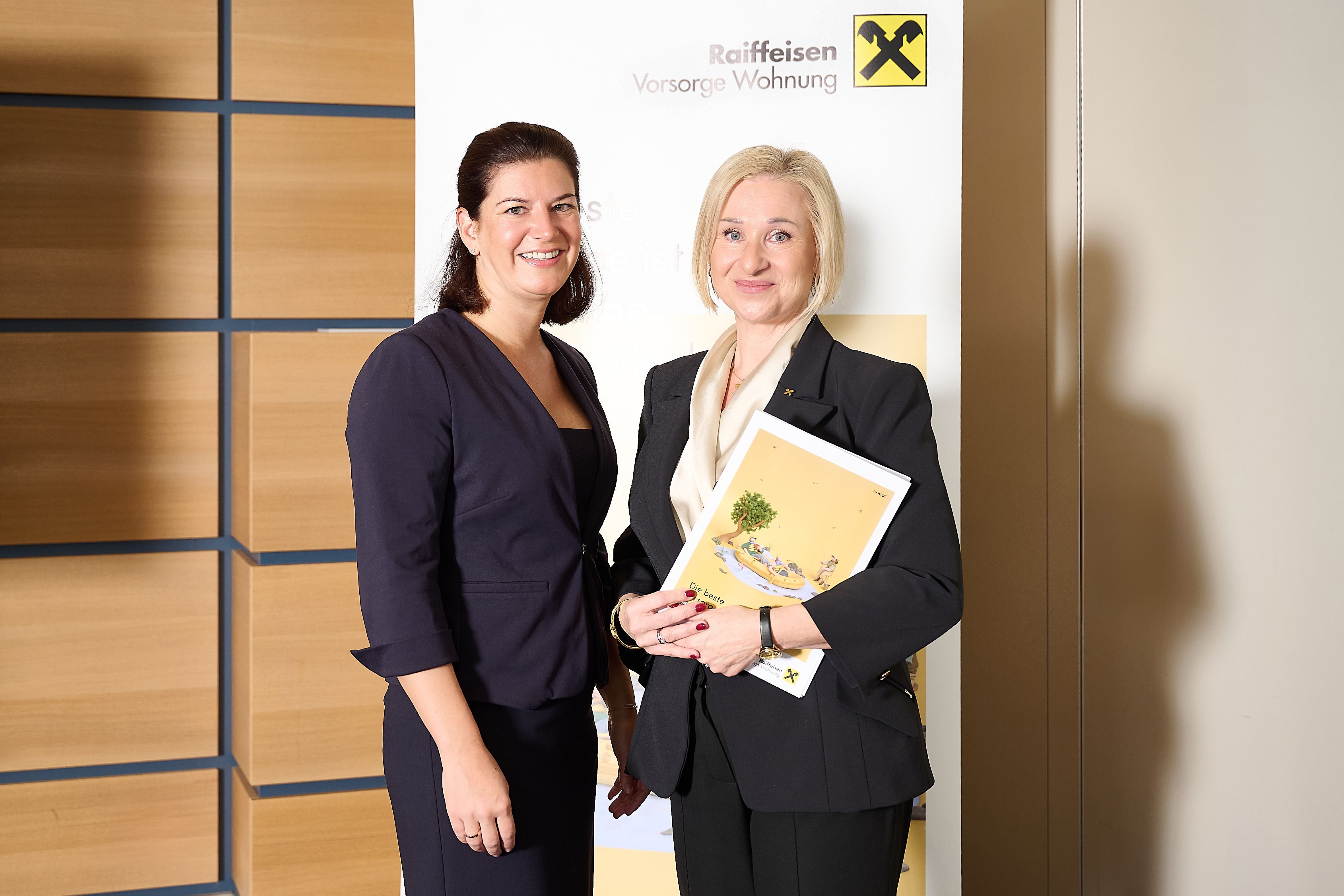 Zwei Frauen in Businesskleidung stehen lächelnd vor einem Raiffeisen-Plakat.