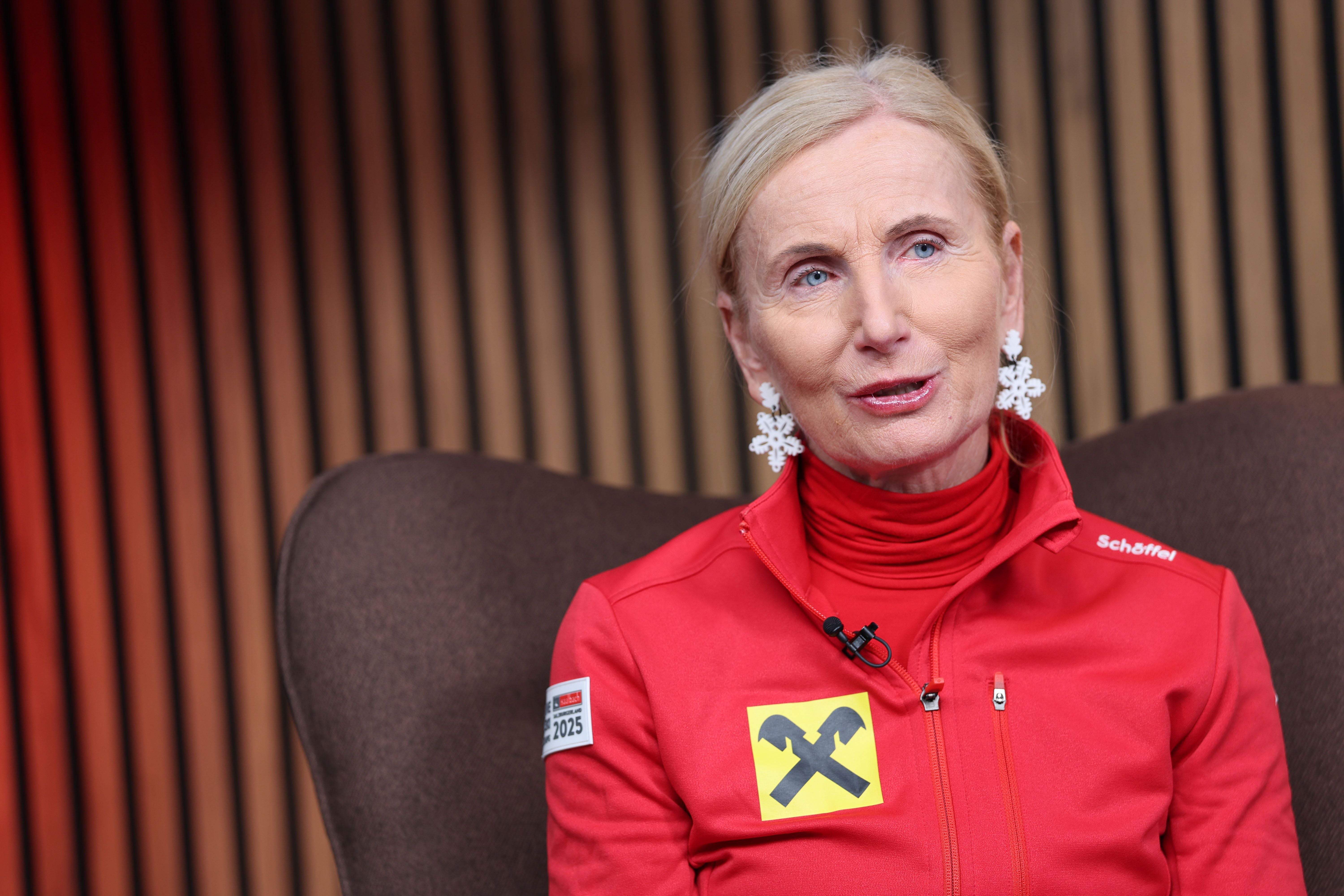 Frau im roten Jackett mit Schneeflockenohrringen sitzt bei einem Interview, Holzlamellen im Hintergrund.