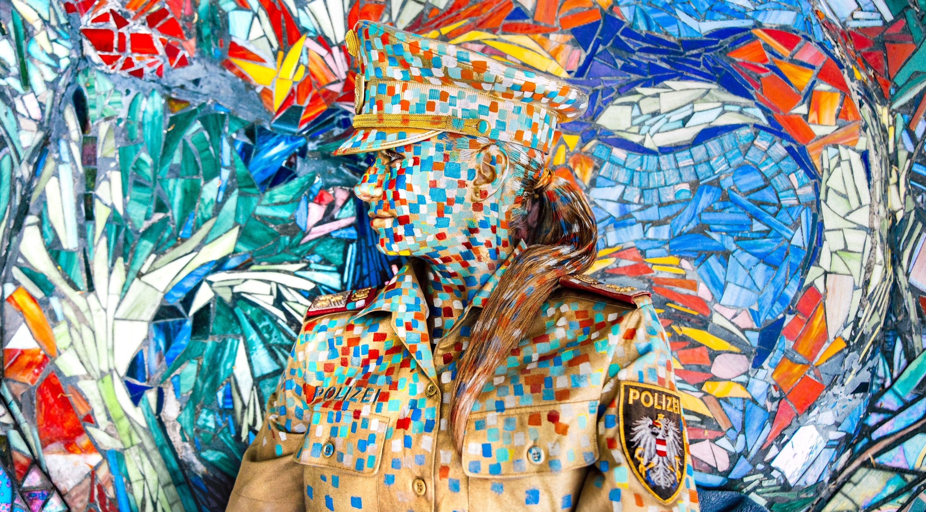 Frau in Polizei-Uniform mit Mosaik-Bodypainting steht vor buntem Mosaik-Hintergrund.