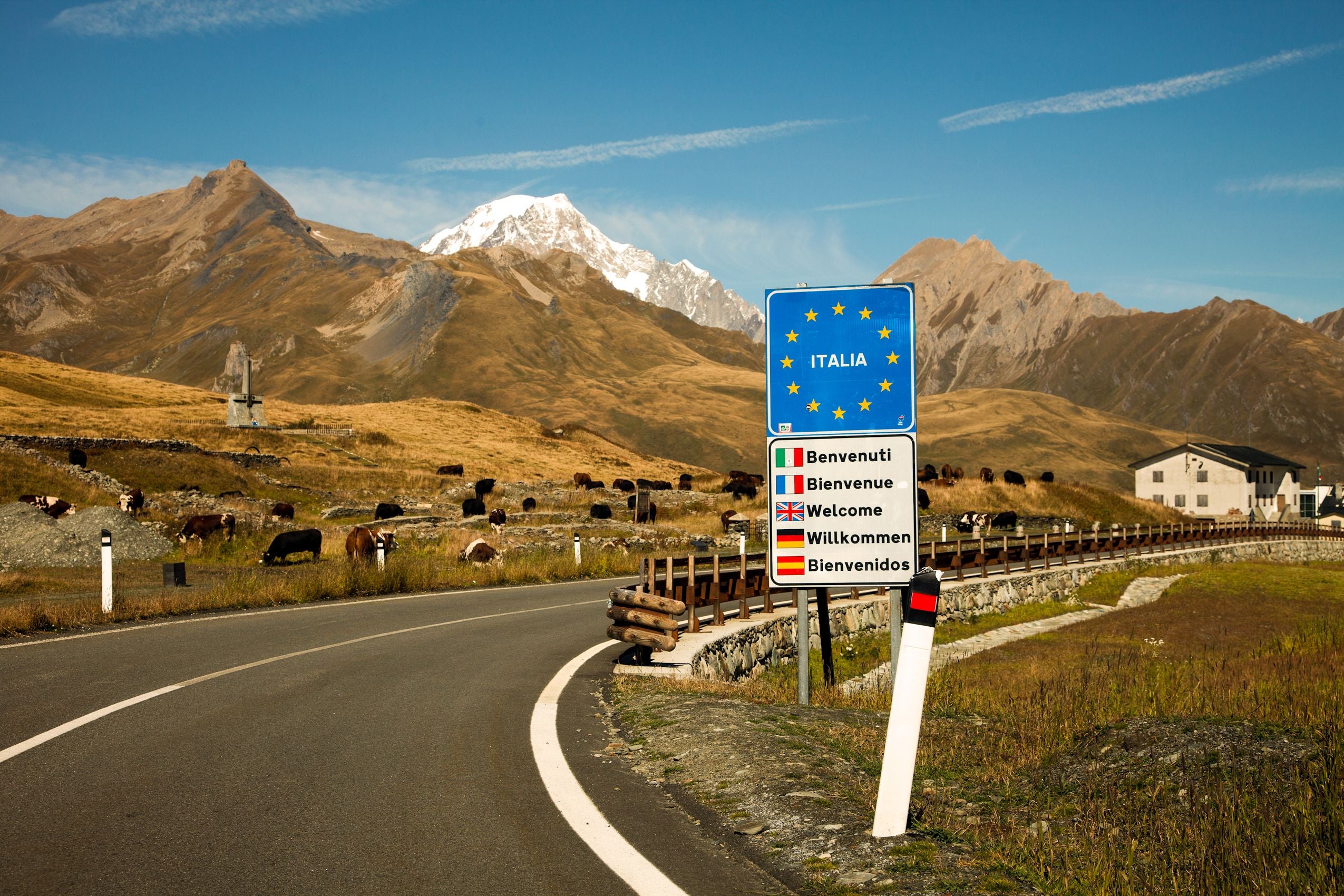 Straßenschild mit Begrüßung an der italienischen Grenze, Berge und Weiden im Hintergrund.