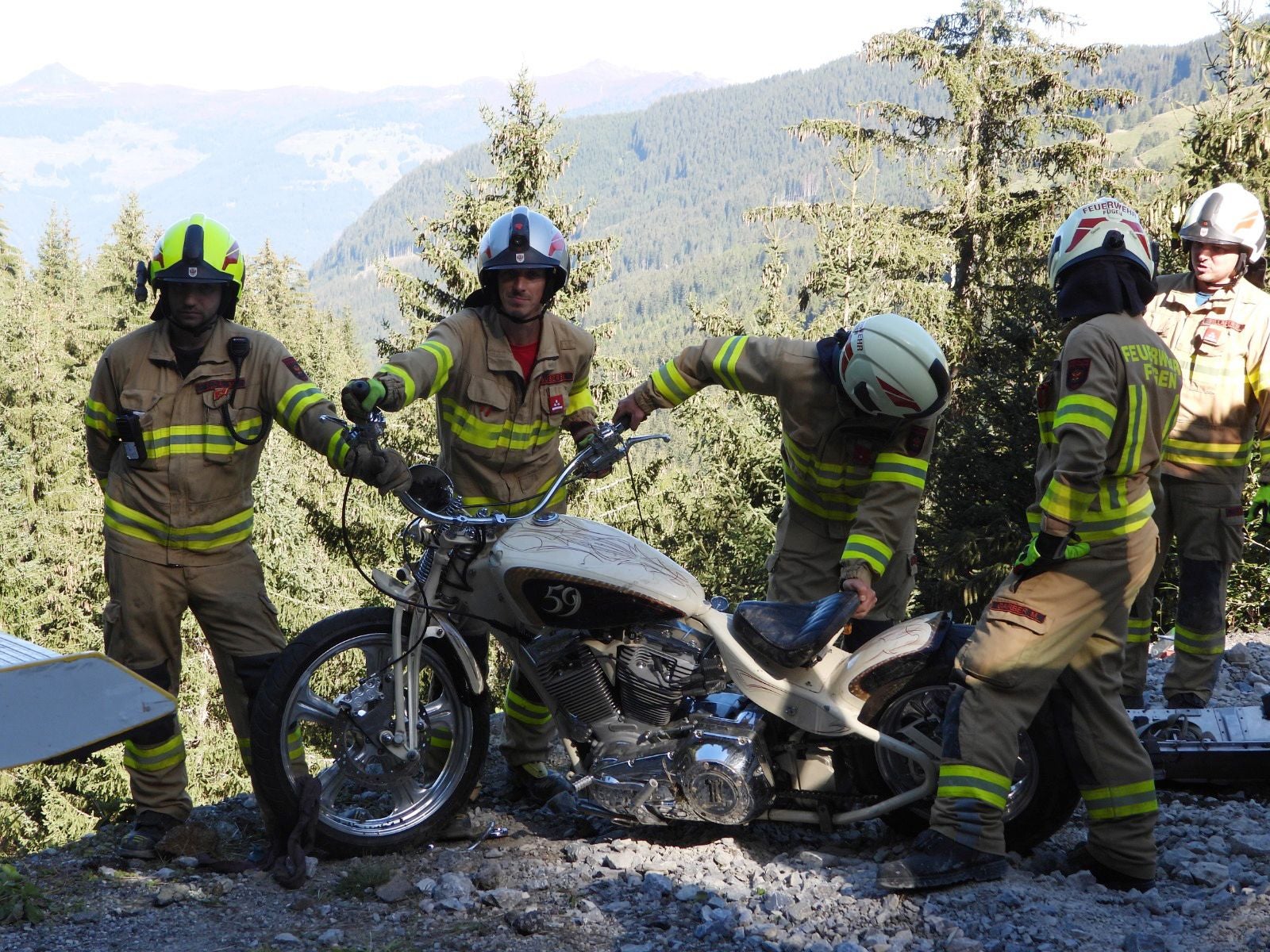 Vier Feuerwehrleute bergen ein Motorrad auf einem steinigen Hang im Gebirge.