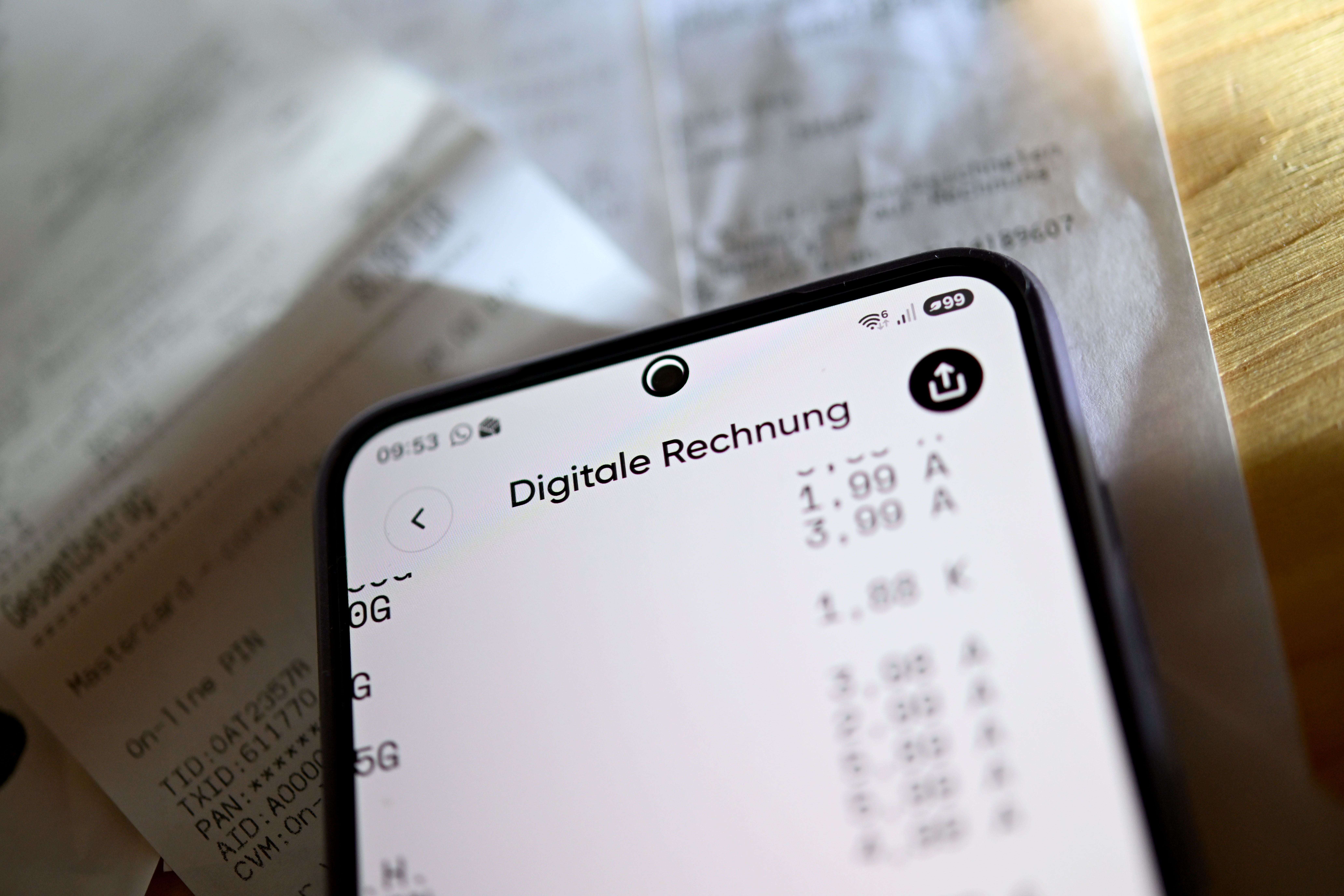 Ein Smartphone zeigt eine digitale Rechnung, darunter liegen unscharfe Papierbelege auf einem Tisch.