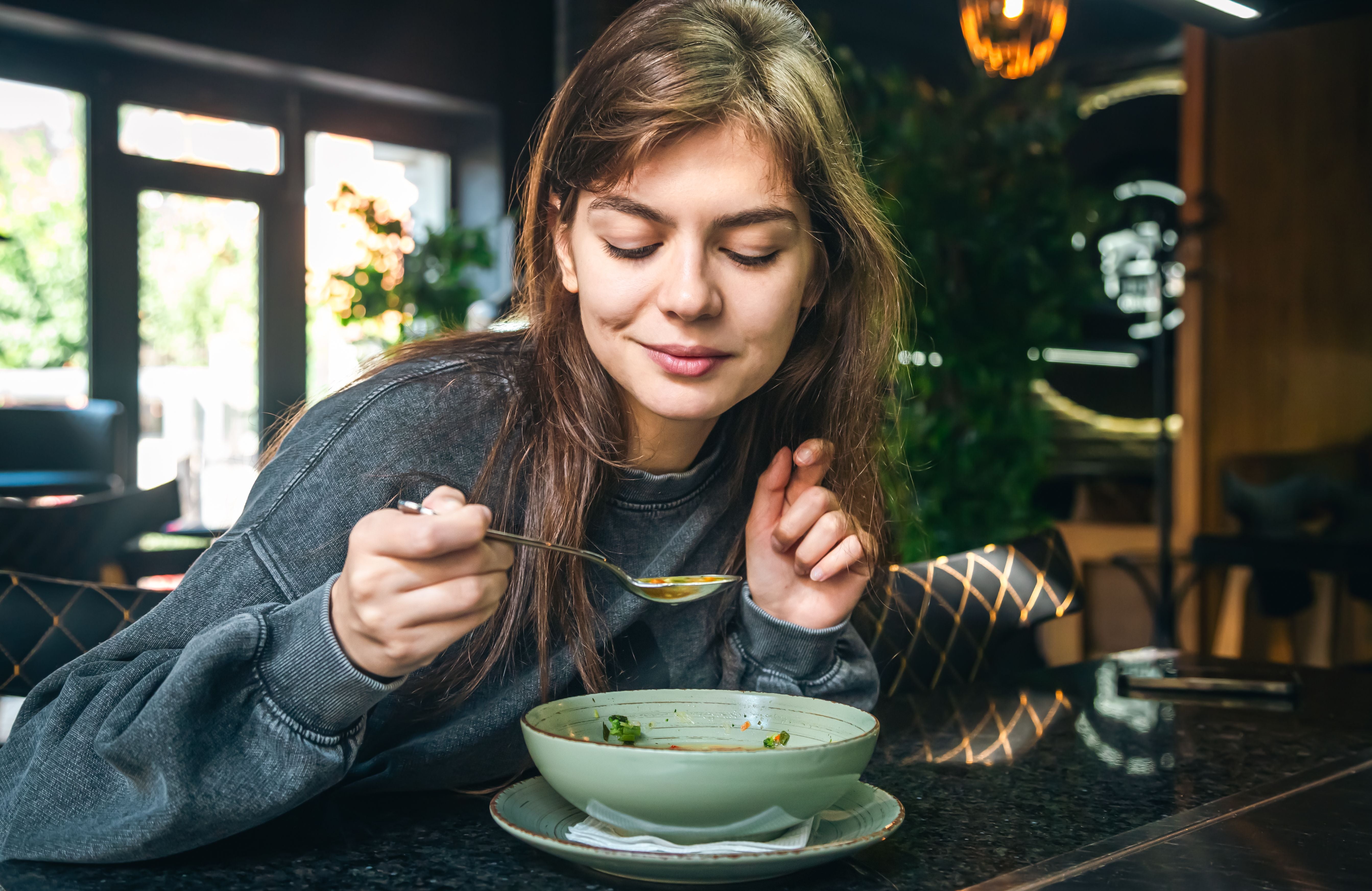Junge Frau isst in einem Café Gemüsesuppe und lächelt zufrieden.