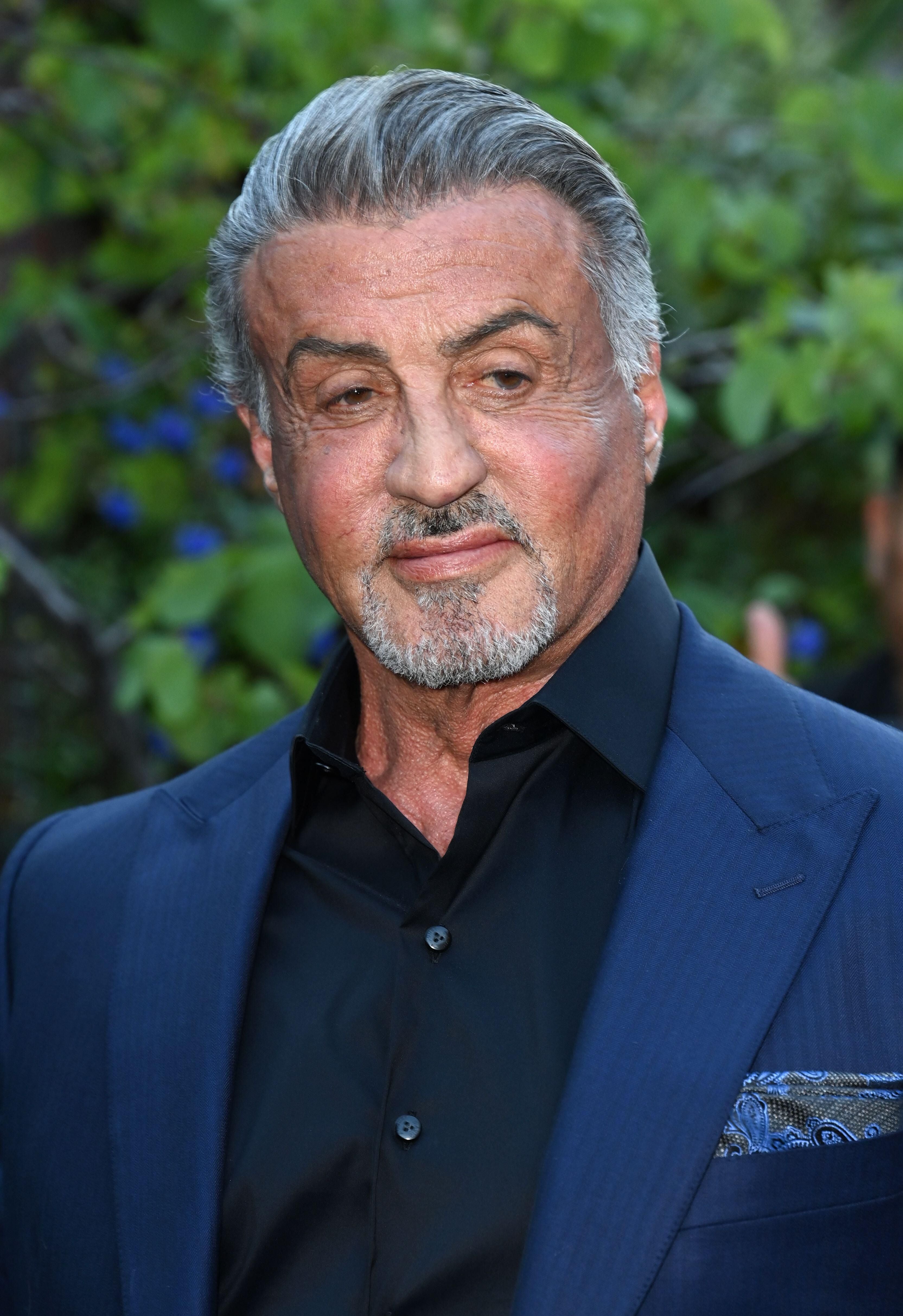 Sylvester Stallone im blauen Anzug und schwarzem Hemd steht im Freien vor grünem Hintergrund.