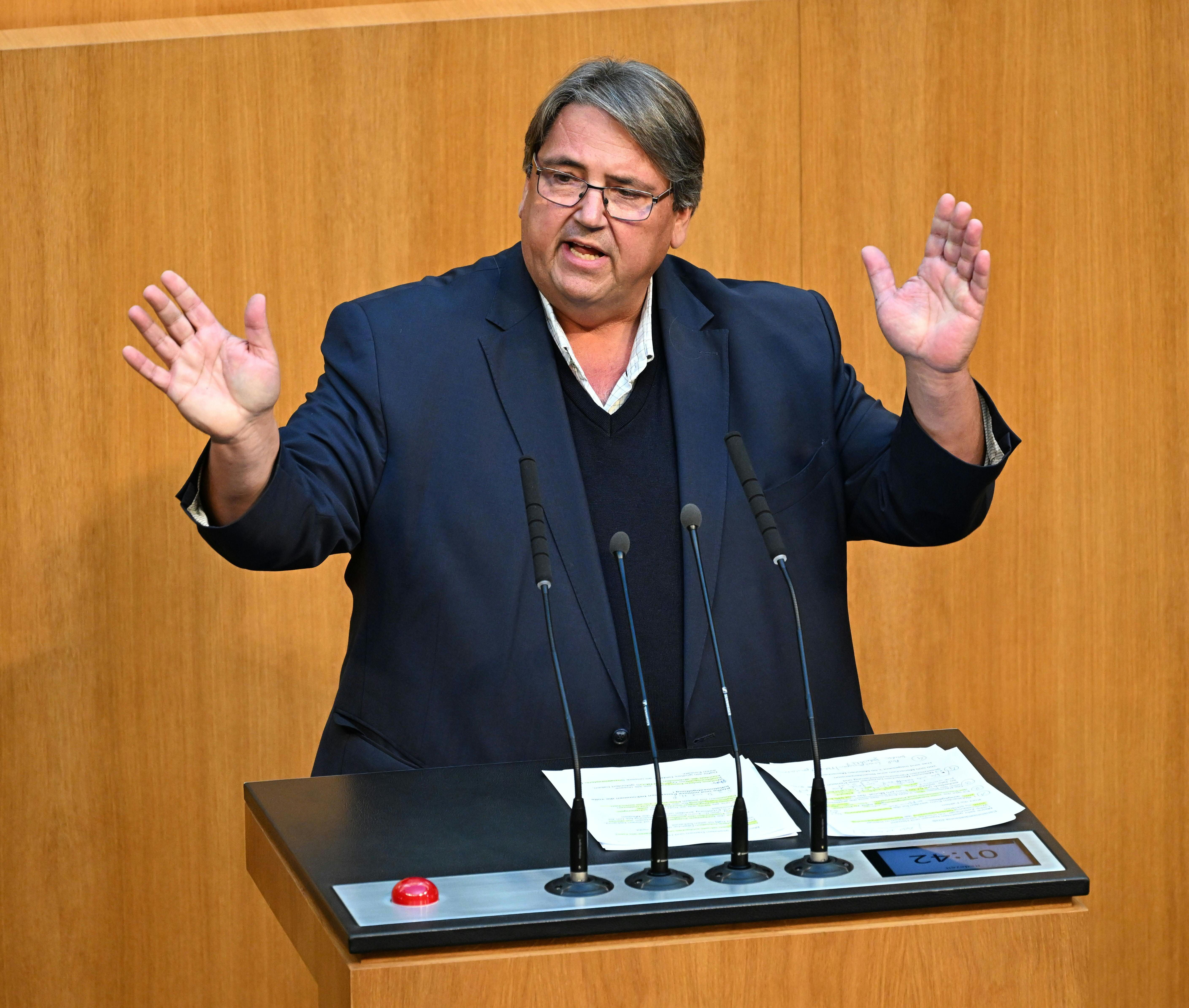 Ein Mann hält eine Rede am Rednerpult in einem Parlamentssaal und gestikuliert mit beiden Händen.