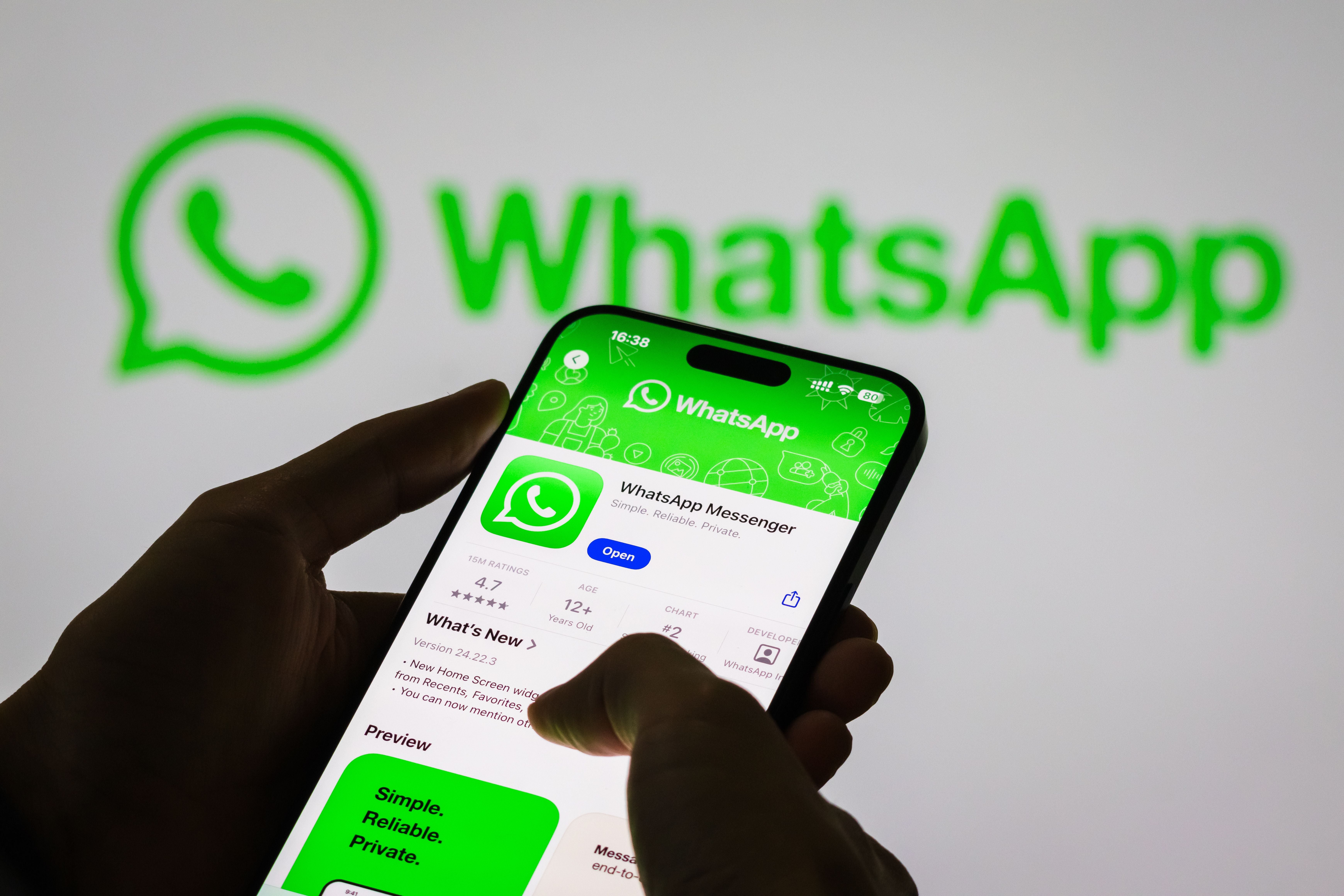 Eine Hand hält ein Smartphone mit geöffneter WhatsApp-App vor großem WhatsApp-Logo im Hintergrund.