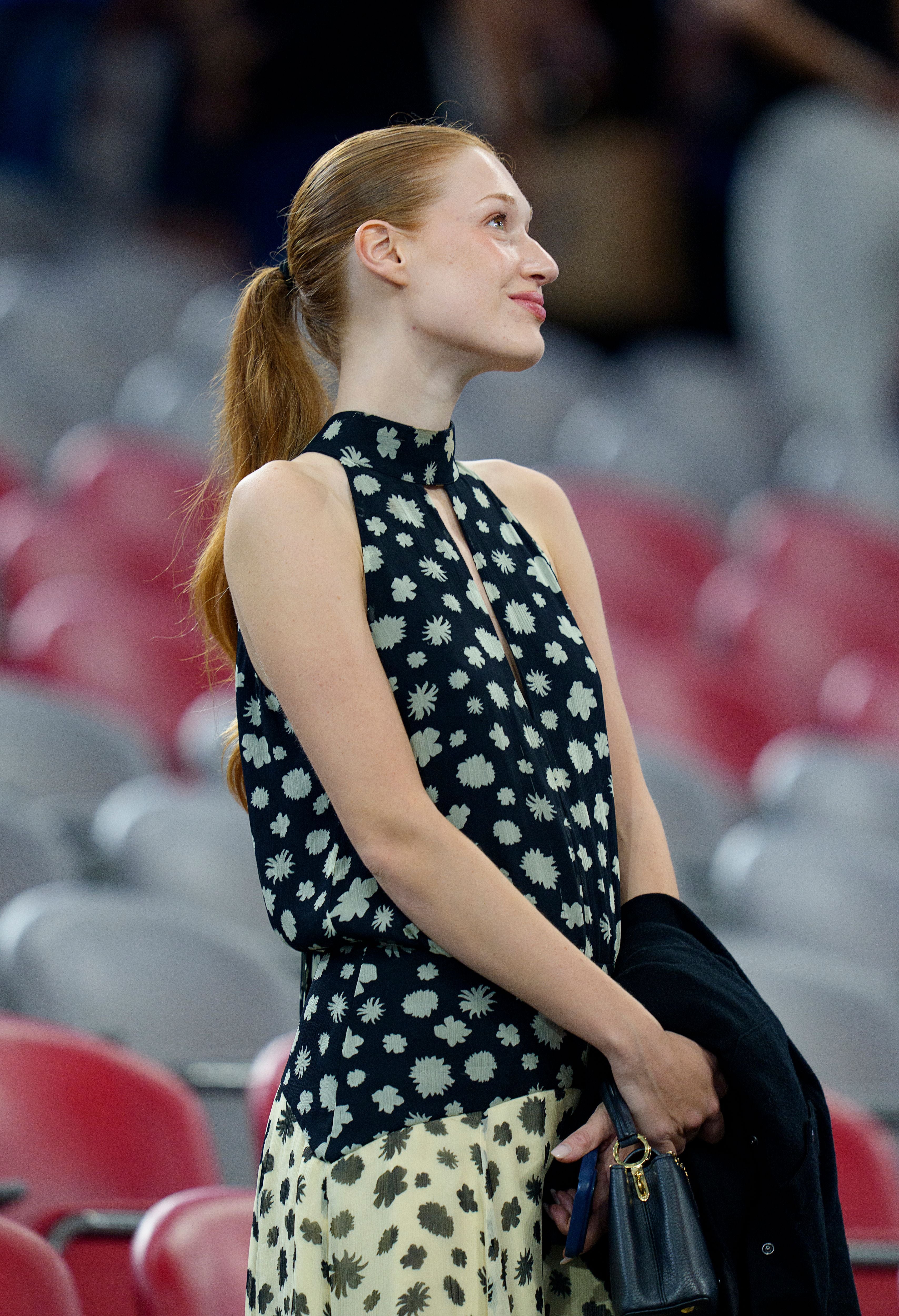 Junge Frau mit blondem Pferdeschwanz steht lächelnd in einem Stadion mit roten und grauen Sitzen.