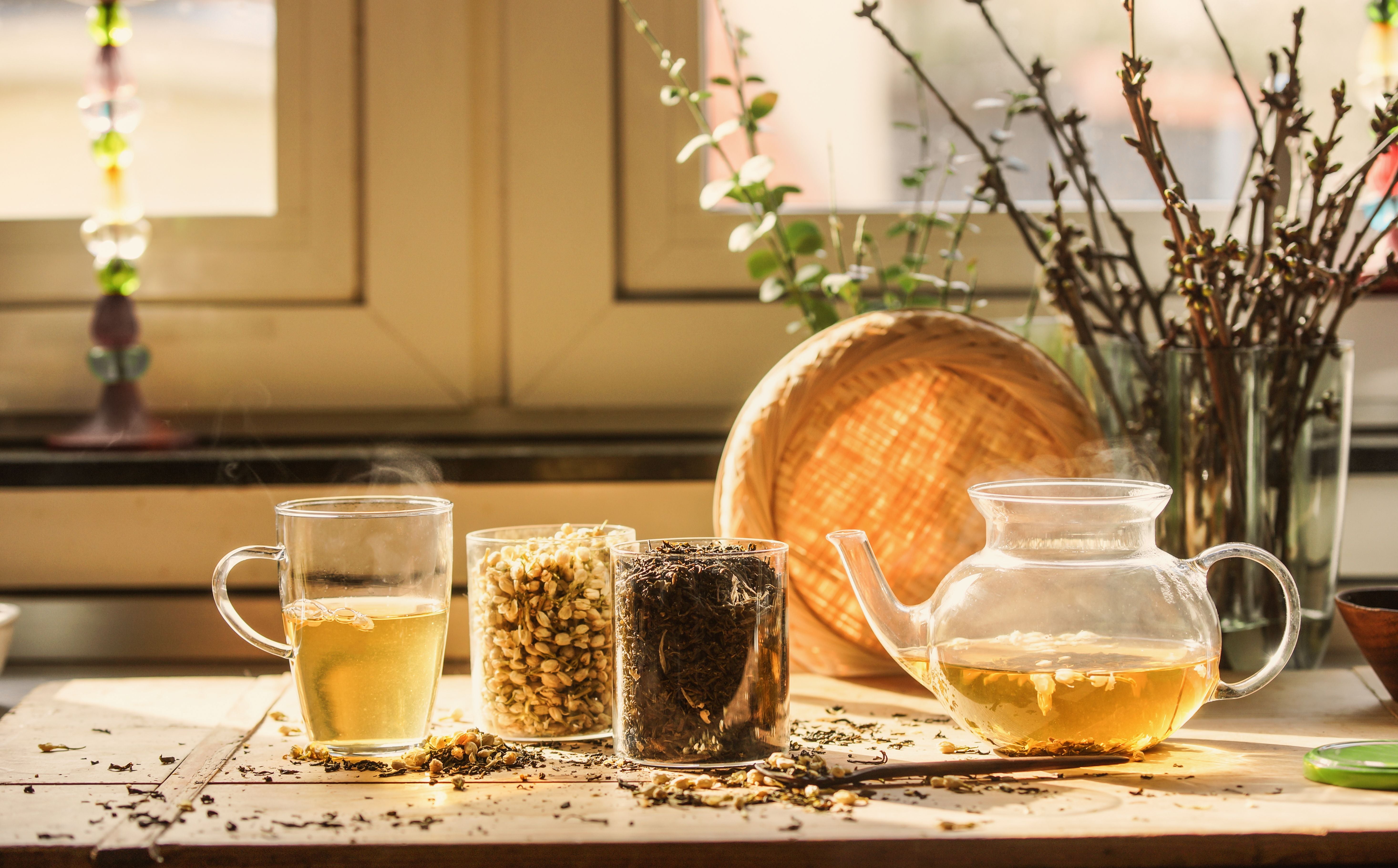Glasbecher und Teekanne mit grünem Jasmintee auf einem sonnigen Küchentisch am Fenster.