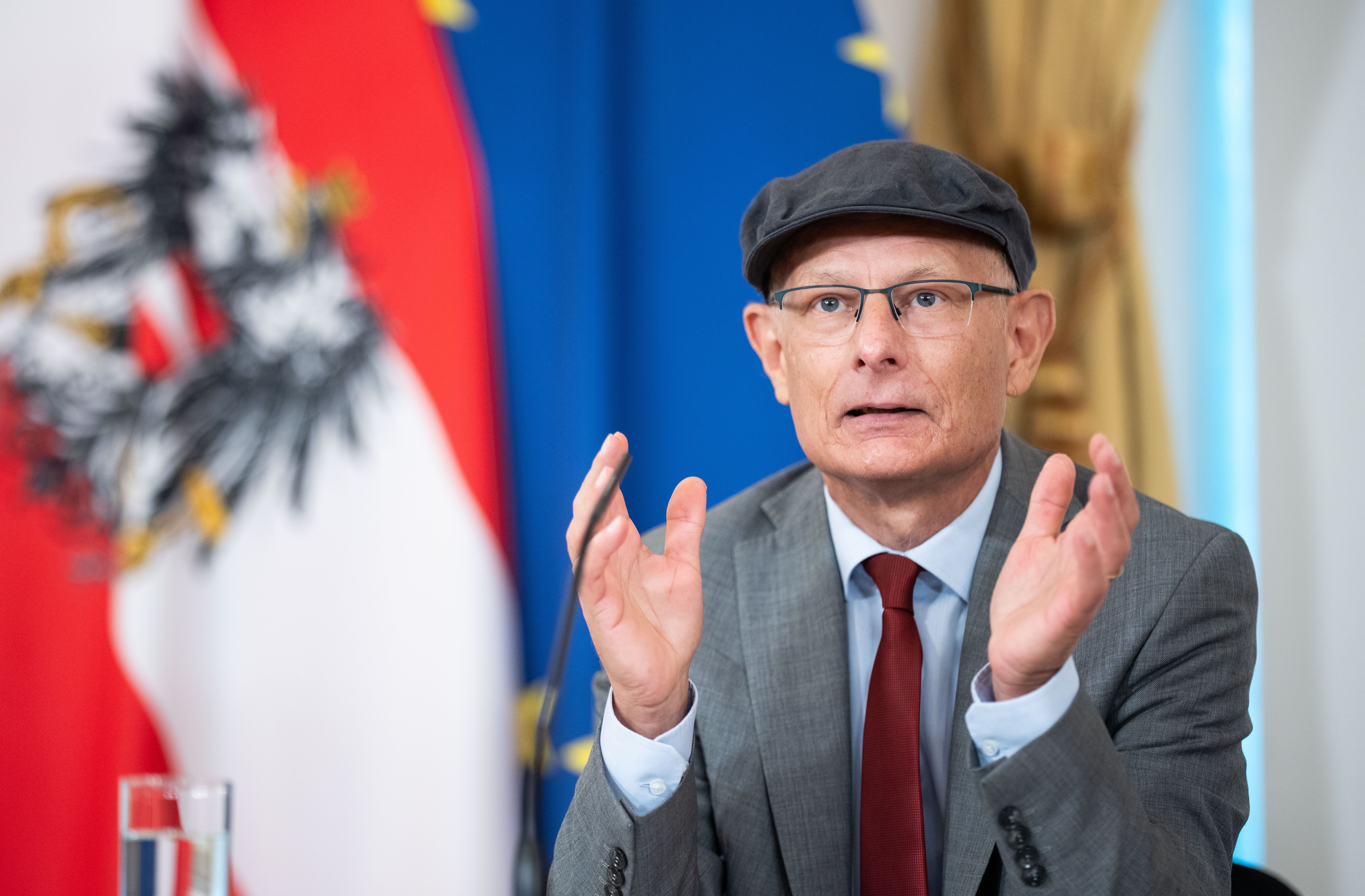 Ein Mann mit Brille und Mütze spricht bei einer Pressekonferenz vor Österreich-Flagge.