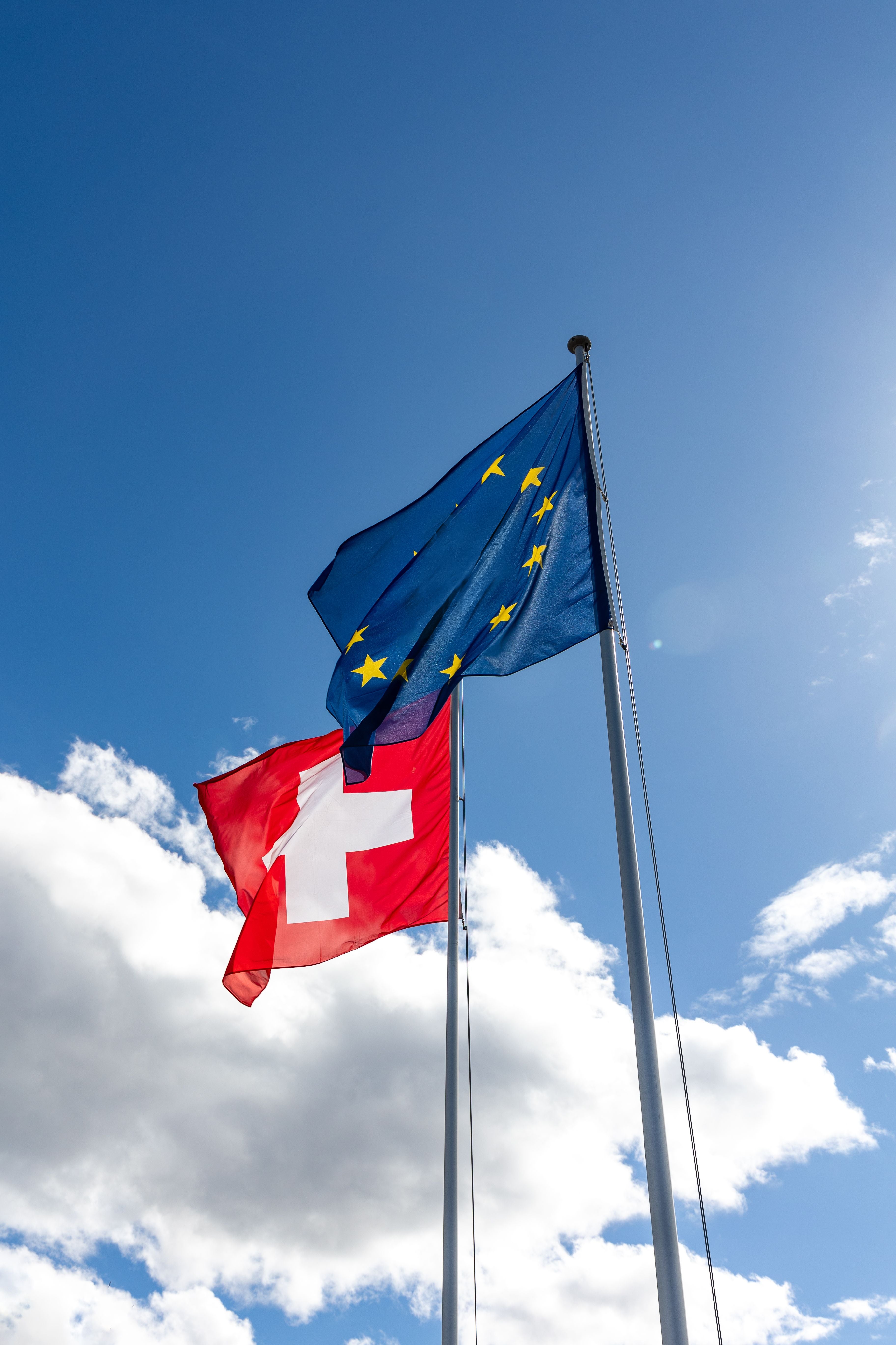 Schweizer und EU-Flagge wehen im Wind vor blauem Himmel mit einigen Wolken.