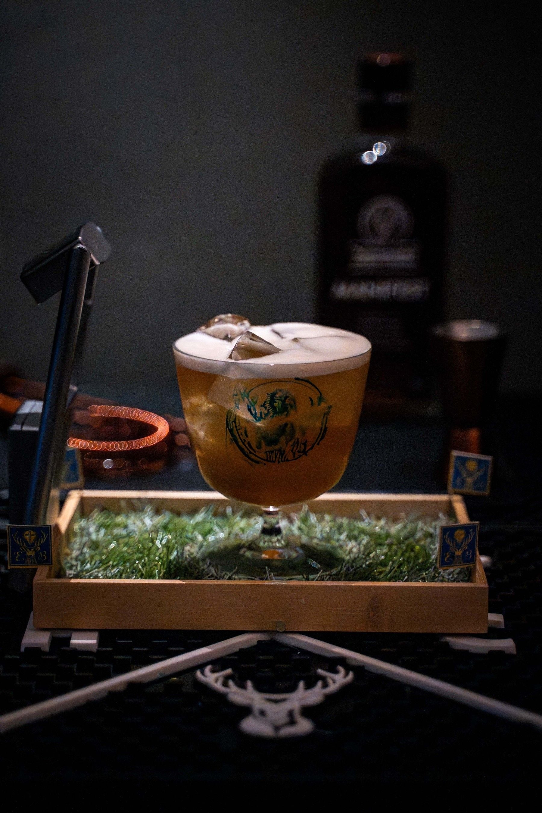 Ein Cocktailglas mit Schaum steht auf einer mit Gras dekorierten Holzplatte, im Hintergrund eine Flasche.
