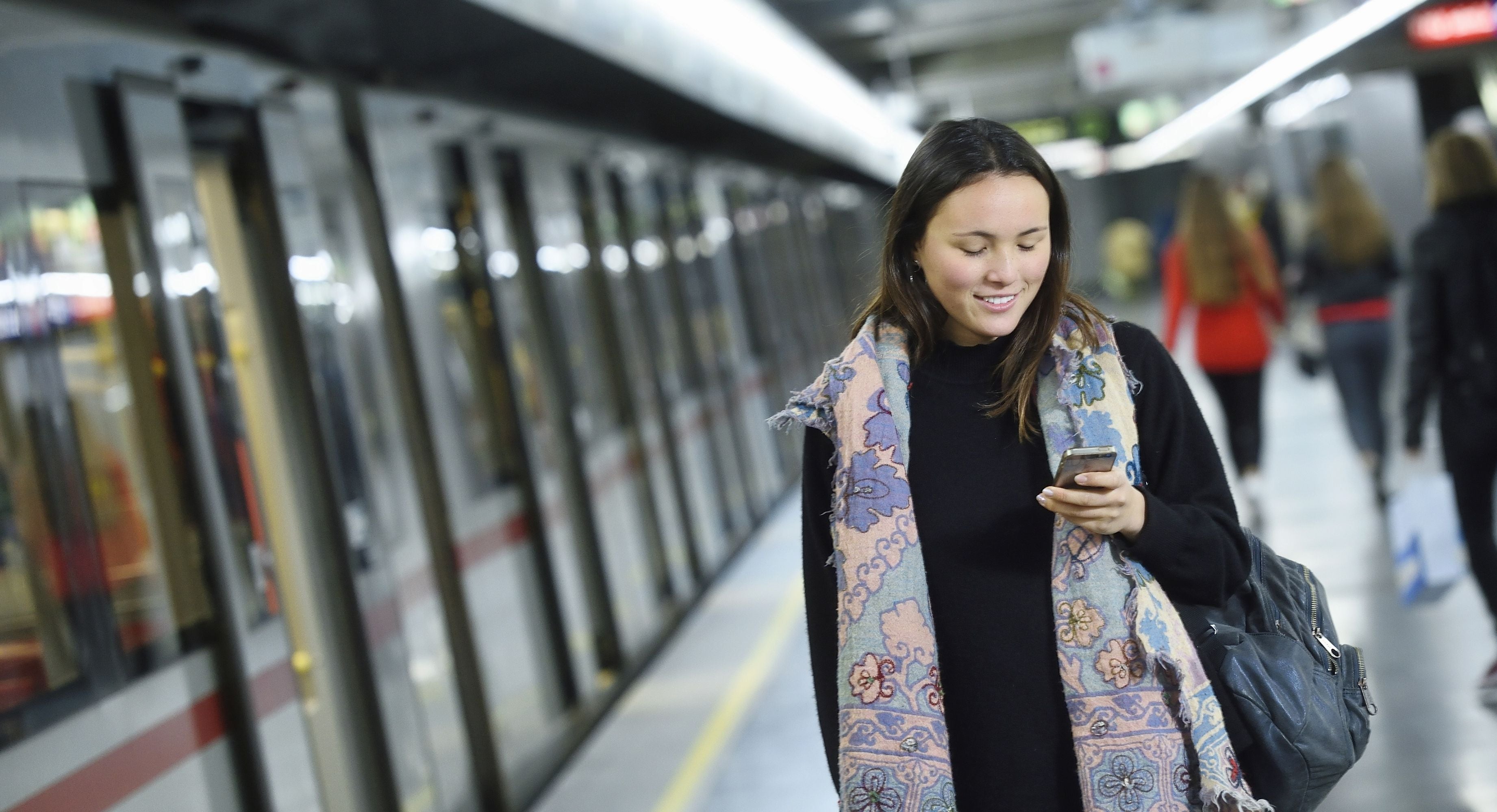 Junge Frau schaut lächelnd auf ihr Smartphone in einer U-Bahn-Station der Wiener Linien.