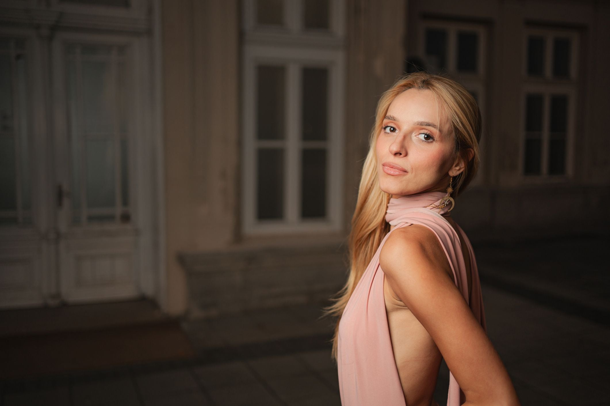 Frau mit langen blonden Haaren posiert selbstbewusst in einem eleganten, rosa Outfit.
