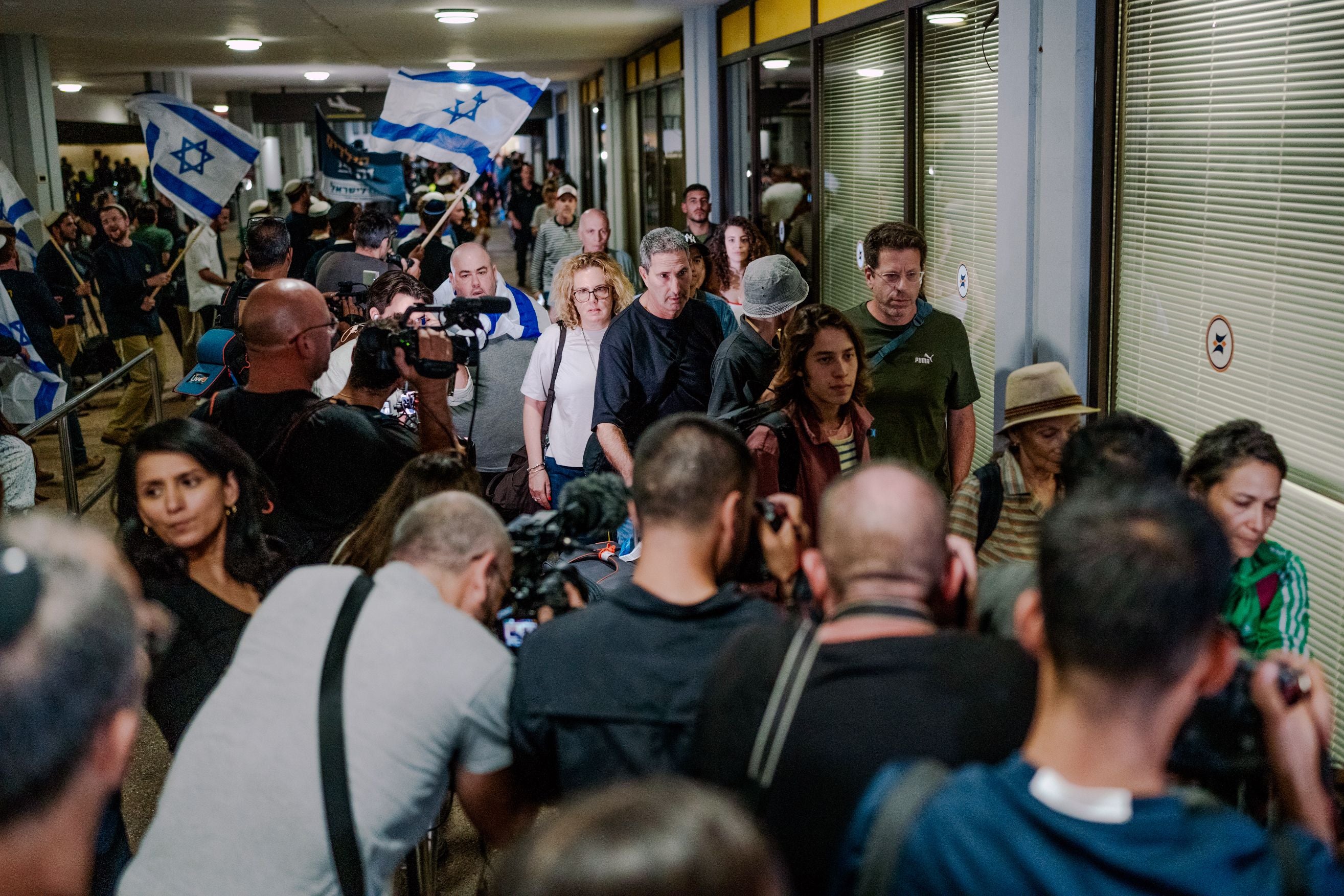 Menschen verlassen einen Flughafen, Fotografen und Reporter warten, israelische Flaggen im Hintergrund.