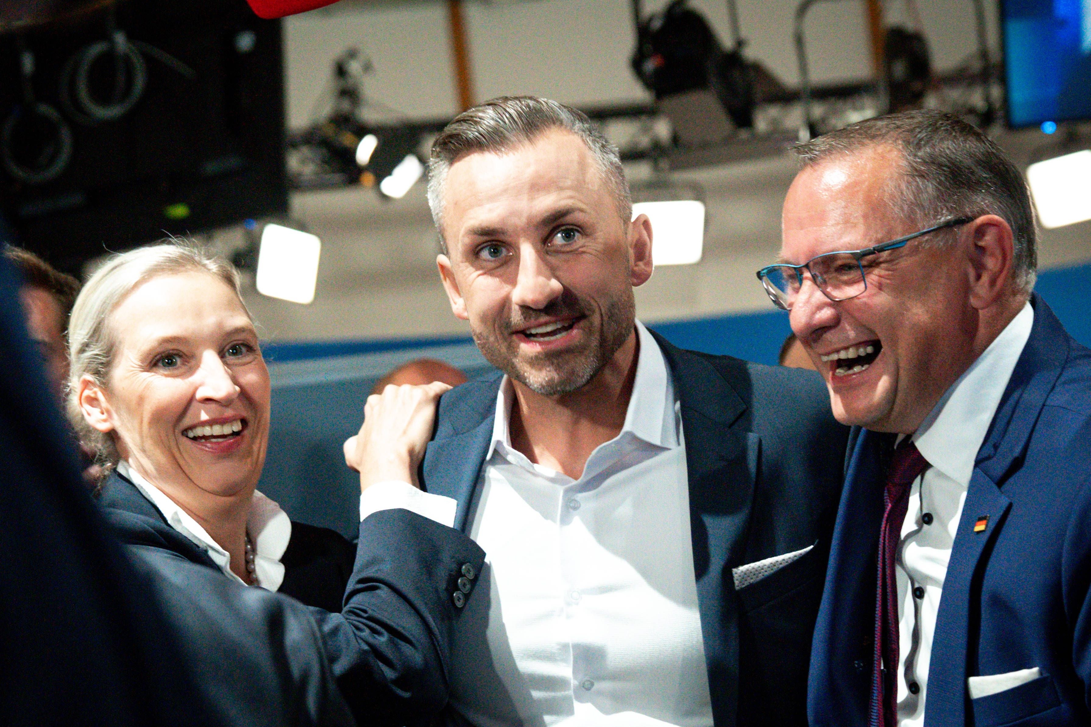 Drei Personen im Anzug jubeln gemeinsam in einer Halle bei einer Wahlveranstaltung.