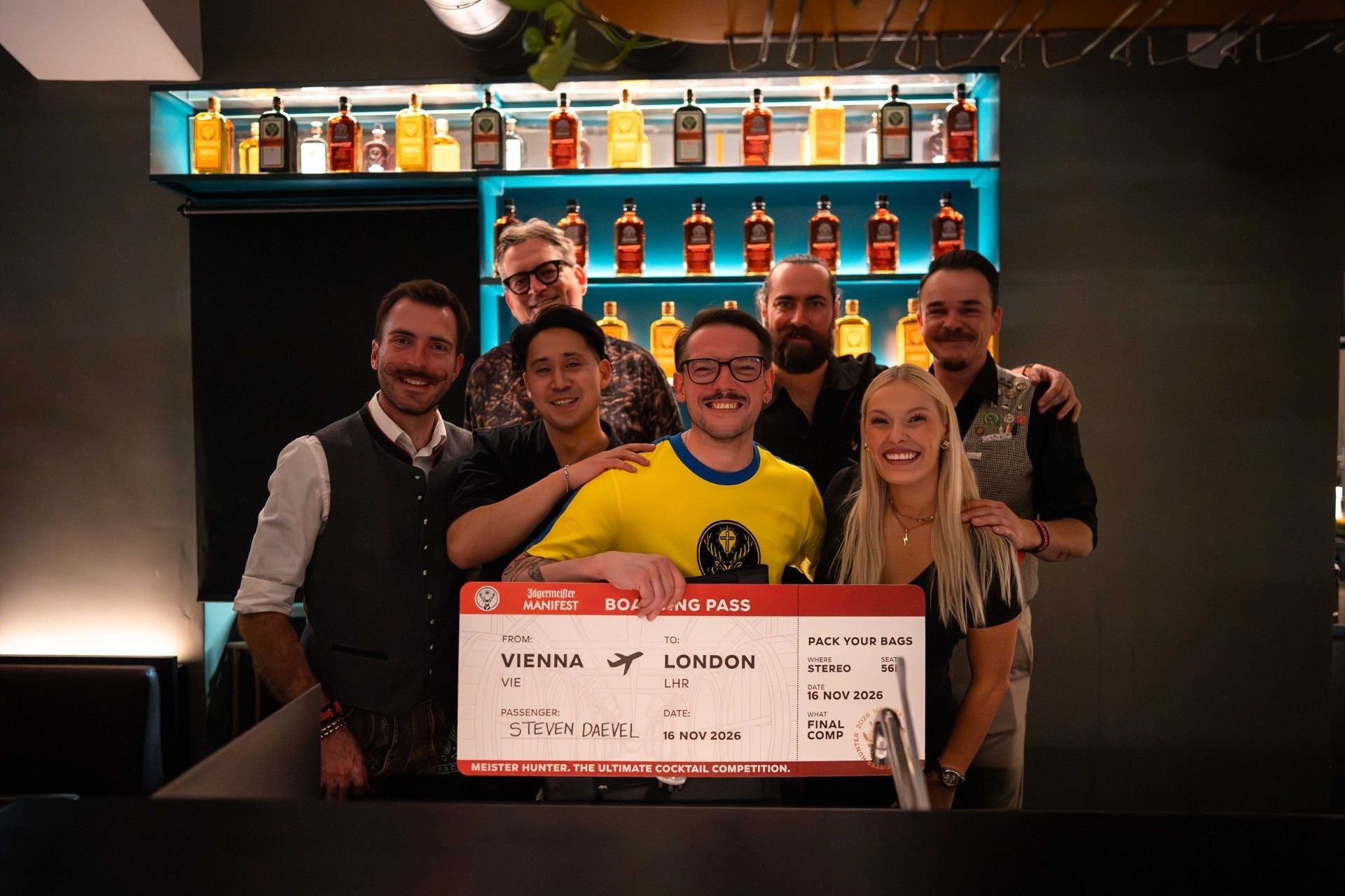 Sieben Menschen posieren lachend in einer Bar mit einer großen Reiseticket-Tafel.