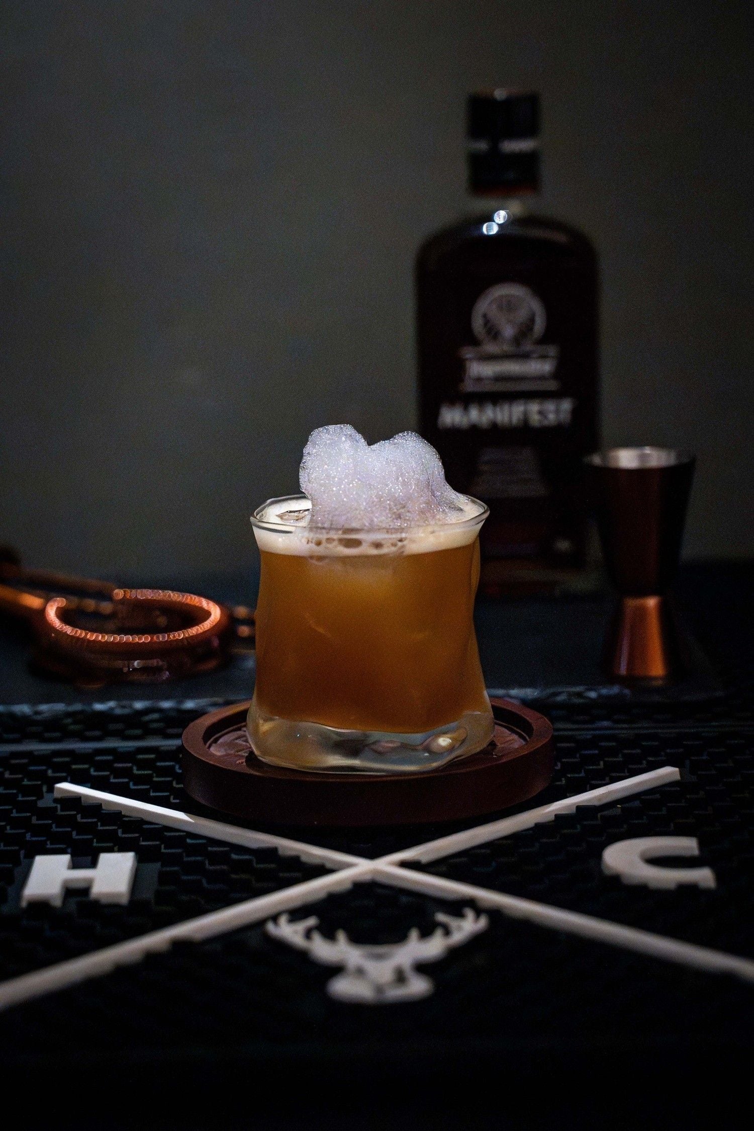 Cocktail mit Schaum in einem Glas auf einem Untersetzer, Flasche im Hintergrund.
