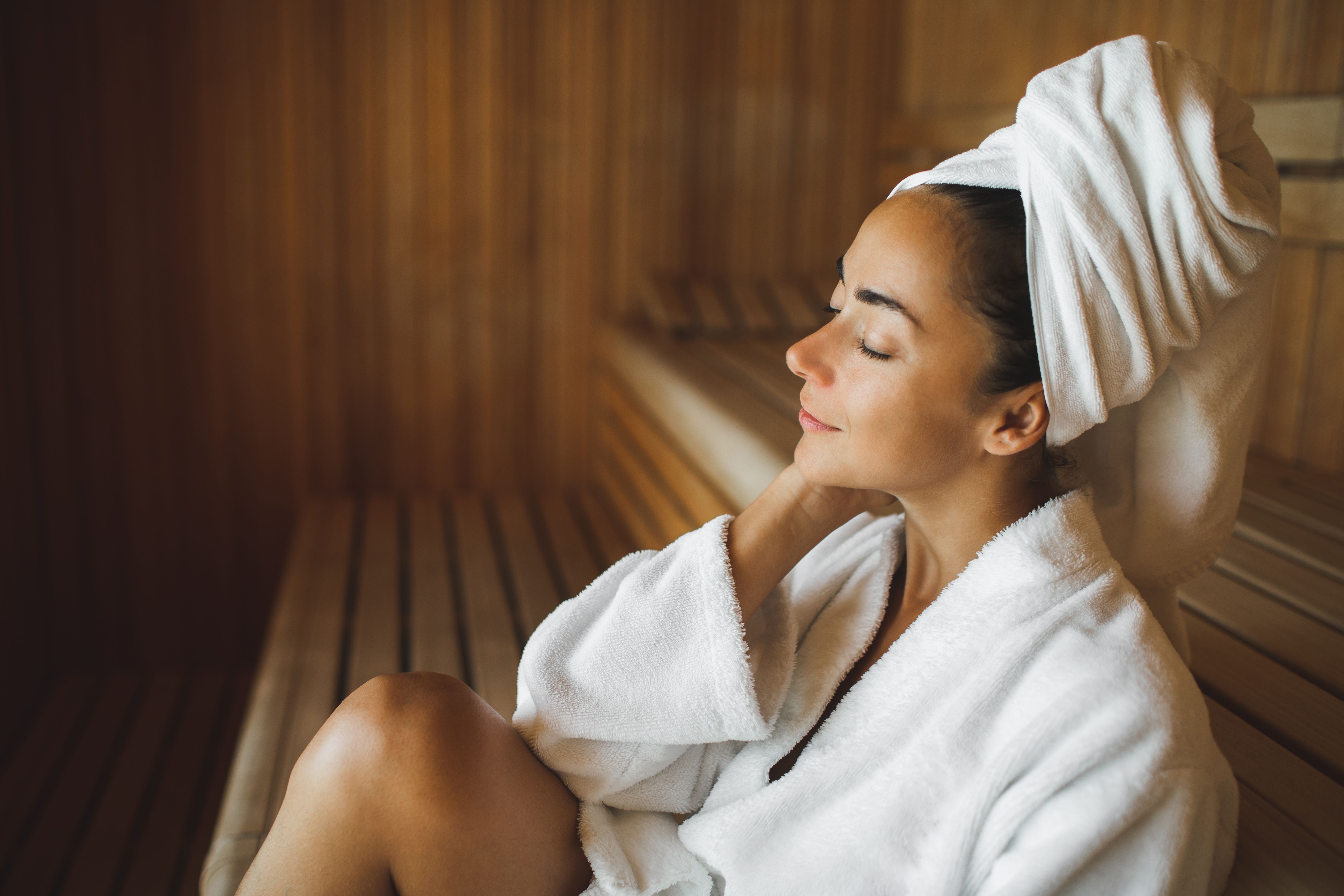 Frau in weißem Bademantel und Handtuchturban entspannt in einer Sauna aus Holz.