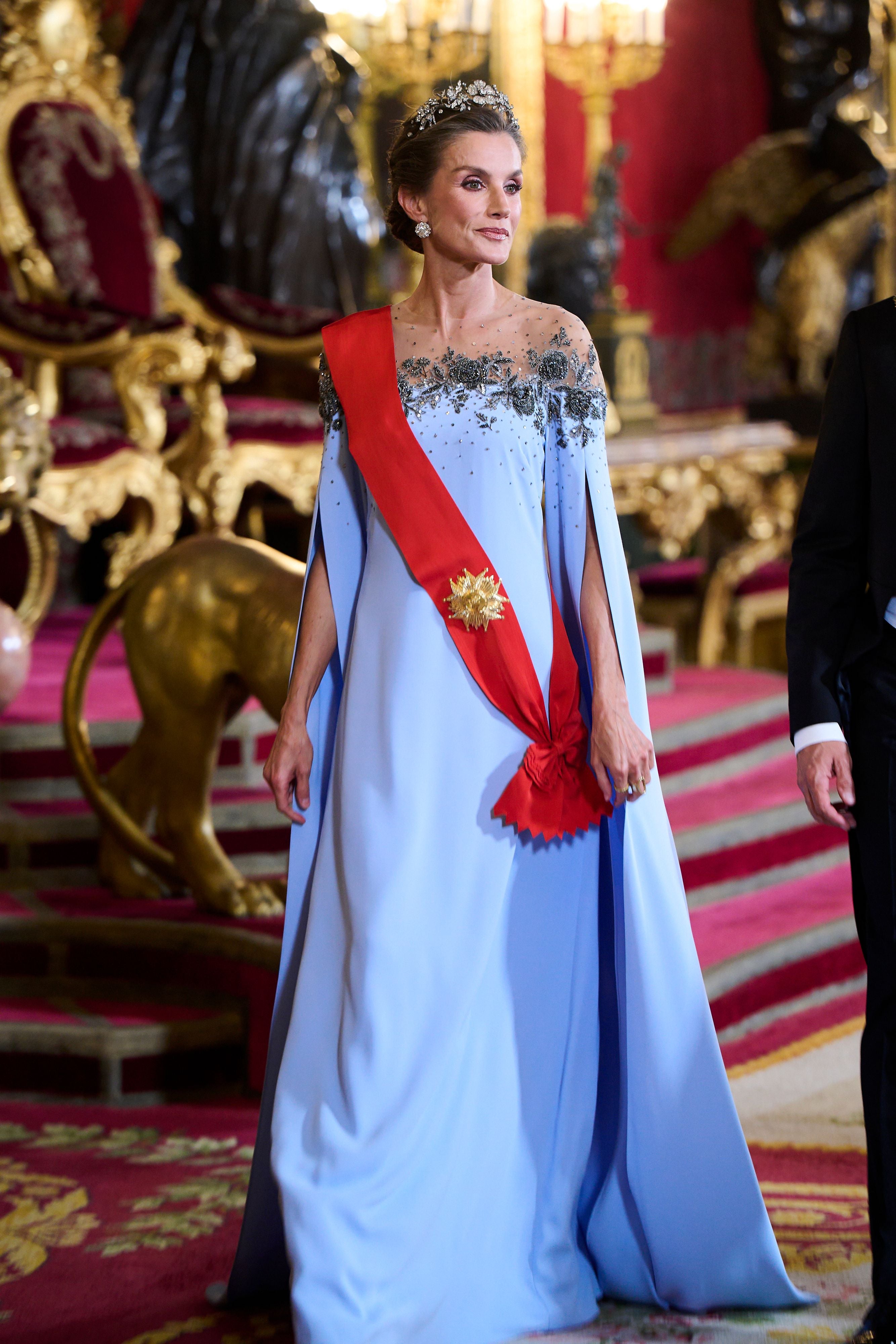 Eine Frau im eleganten, blauen Kleid mit rotem Band steht in prunkvollem, goldverzierten Palast.