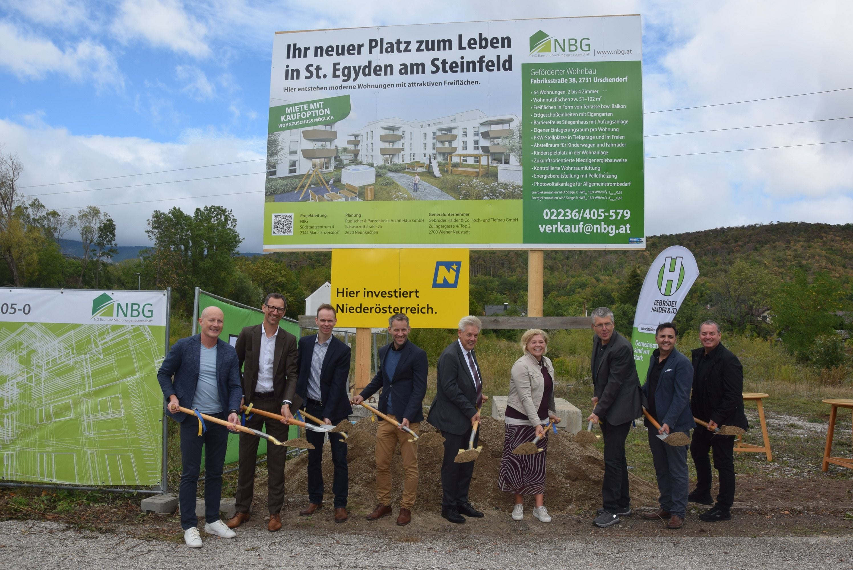 Neun Personen mit Spaten beim Spatenstich vor einem Bauschild und Werbetafeln für ein Wohnprojekt.