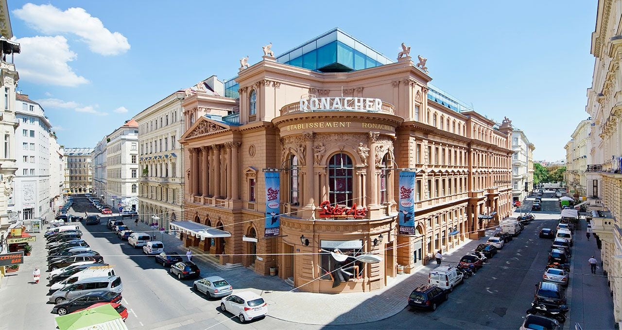 Das Ronacher Theater in Wien an einer Straßenecke bei sonnigem Wetter, Autos parken ringsum.