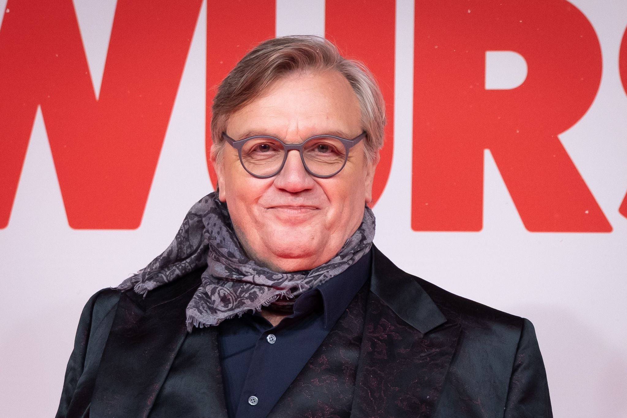 Ein Mann mit Brille und Schal lächelt vor einem rot-weißen Hintergrund.