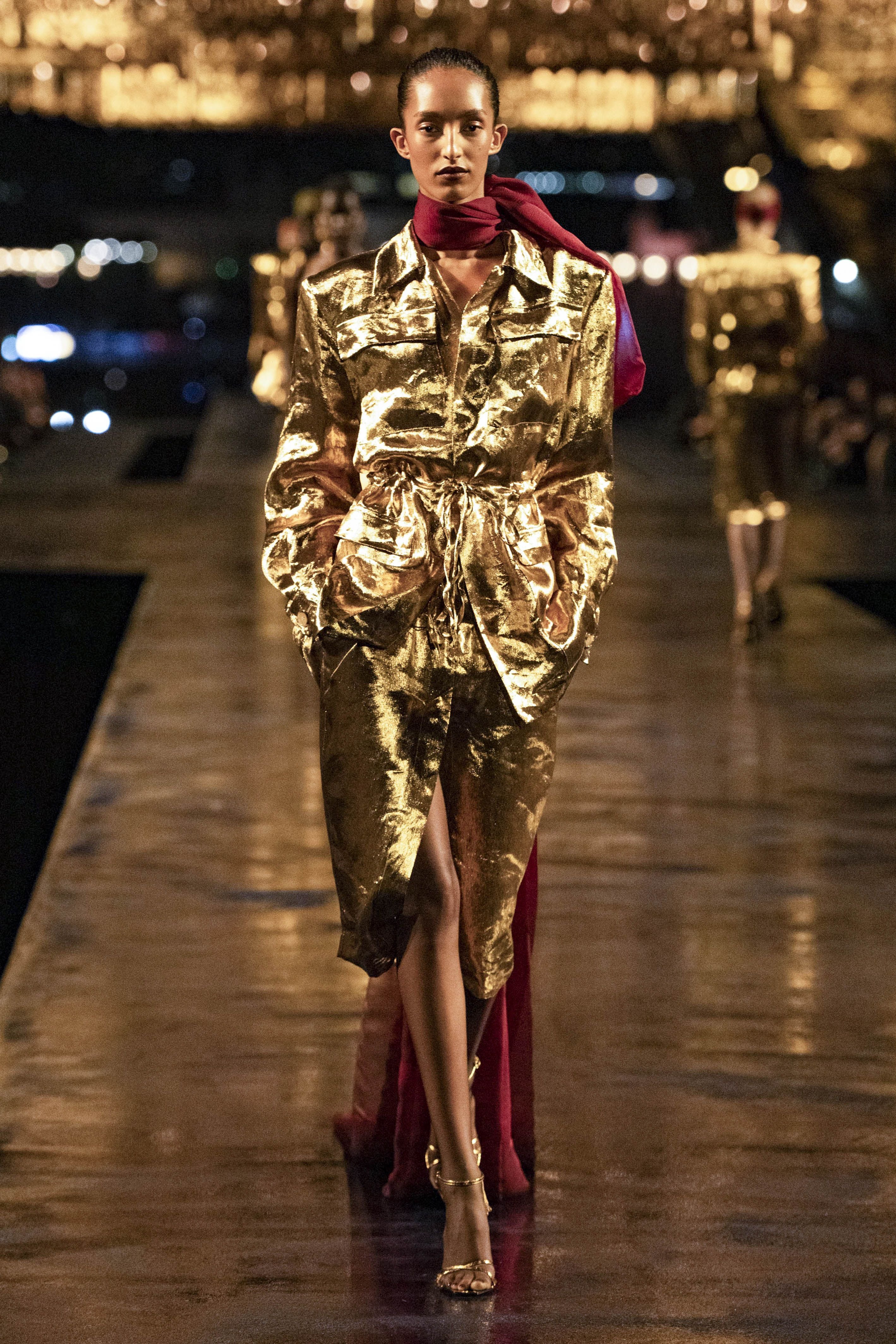 Model trägt goldenen Glanzmantel und roten Schal auf dem Laufsteg bei einer Modenschau.